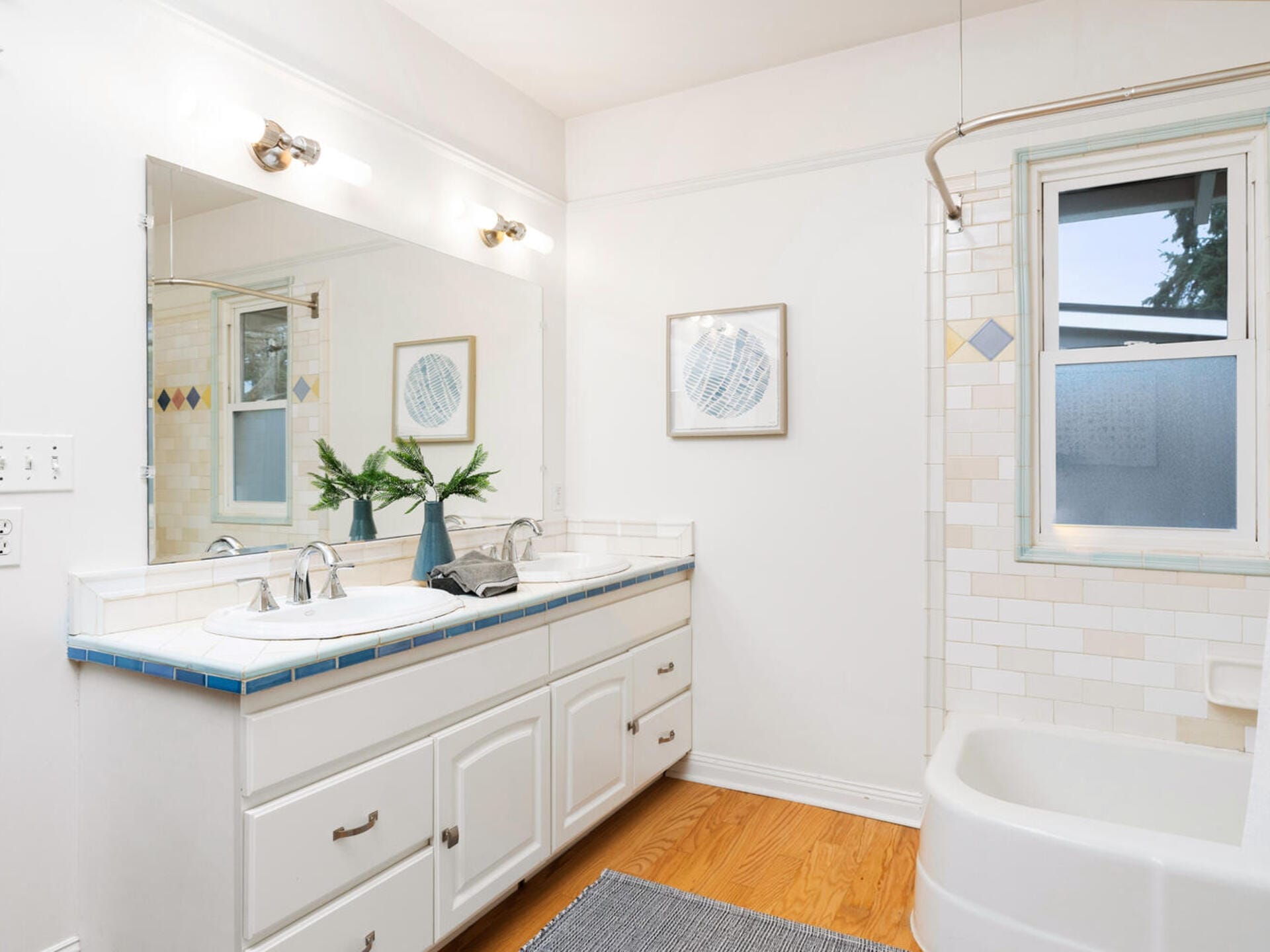 A bright bathroom with a dual-sink vanity featuring a large mirror above it. The vanity has white cabinets and a vase with greenery. The room is illuminated by wall-mounted lights and has a bathtub with light-colored tiles and a window.