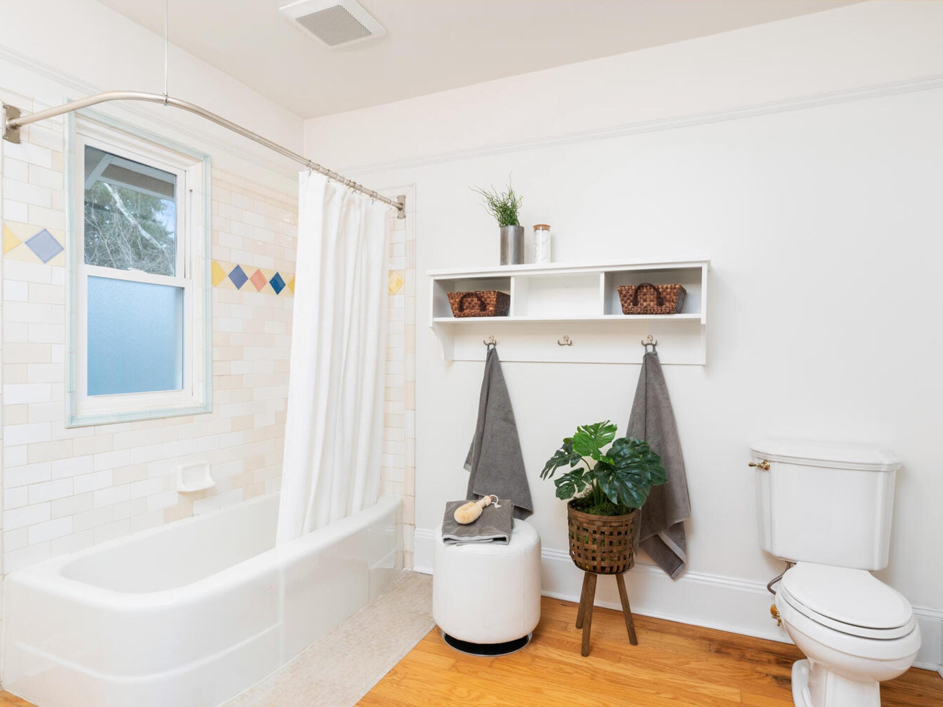 A bright bathroom with a white bathtub and curtain, a window, and a white toilet. There are colorful tiles above the tub, a white shelf with decor, two gray towels hanging, a plant on a stool, and a wooden floor.