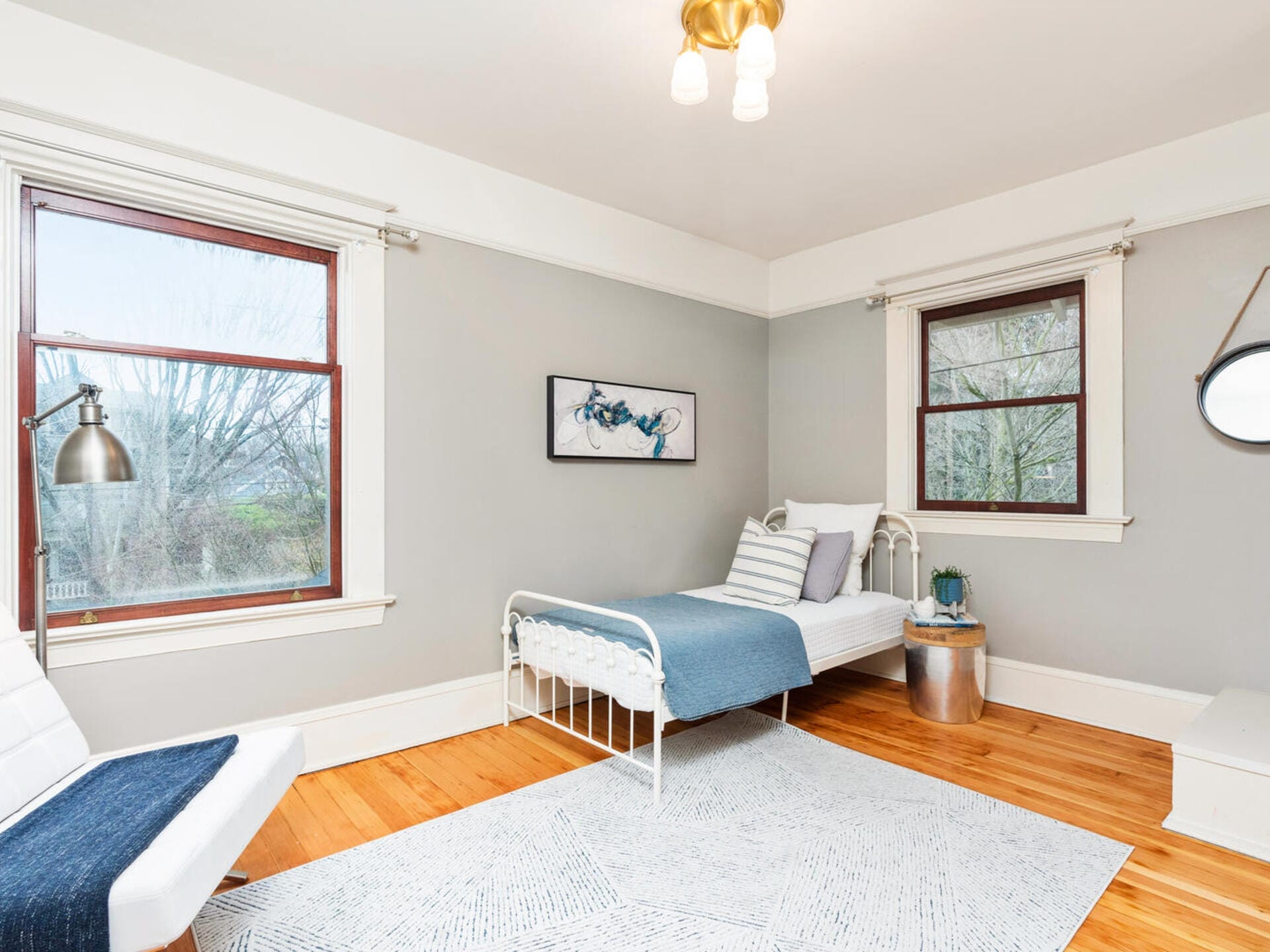 A minimalist bedroom with a single metal bed featuring blue and white bedding. A small round table holds a plant. A large window offers natural light, and a modern ceiling light hangs above. A light gray rug covers the wooden floor.
