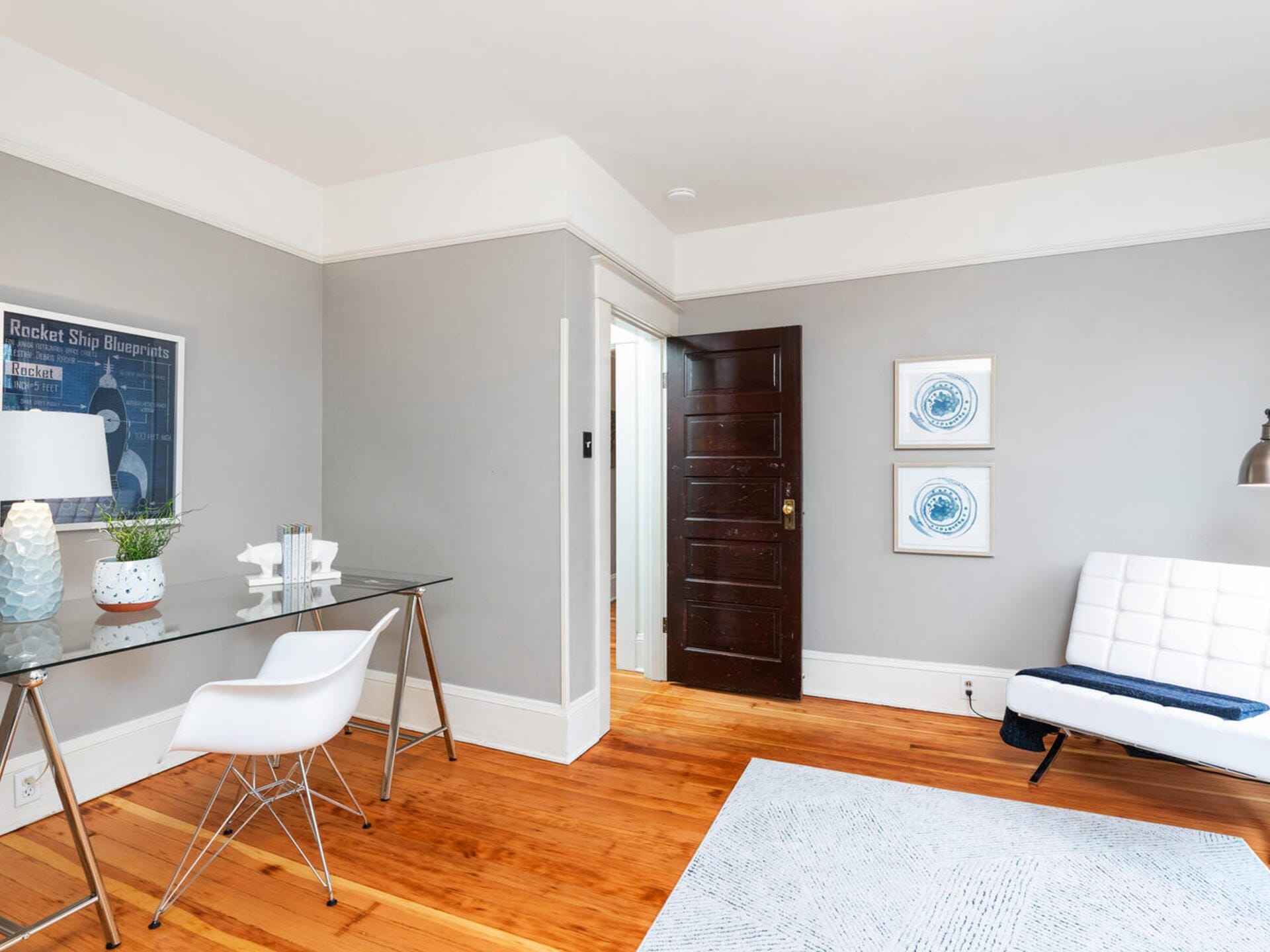 A modern, minimalist living room with light gray walls, a wooden floor, and a white sofa. Theres a glass desk with a white chair, a table lamp, and decorative items. Two framed artworks and a dark wooden door are visible.