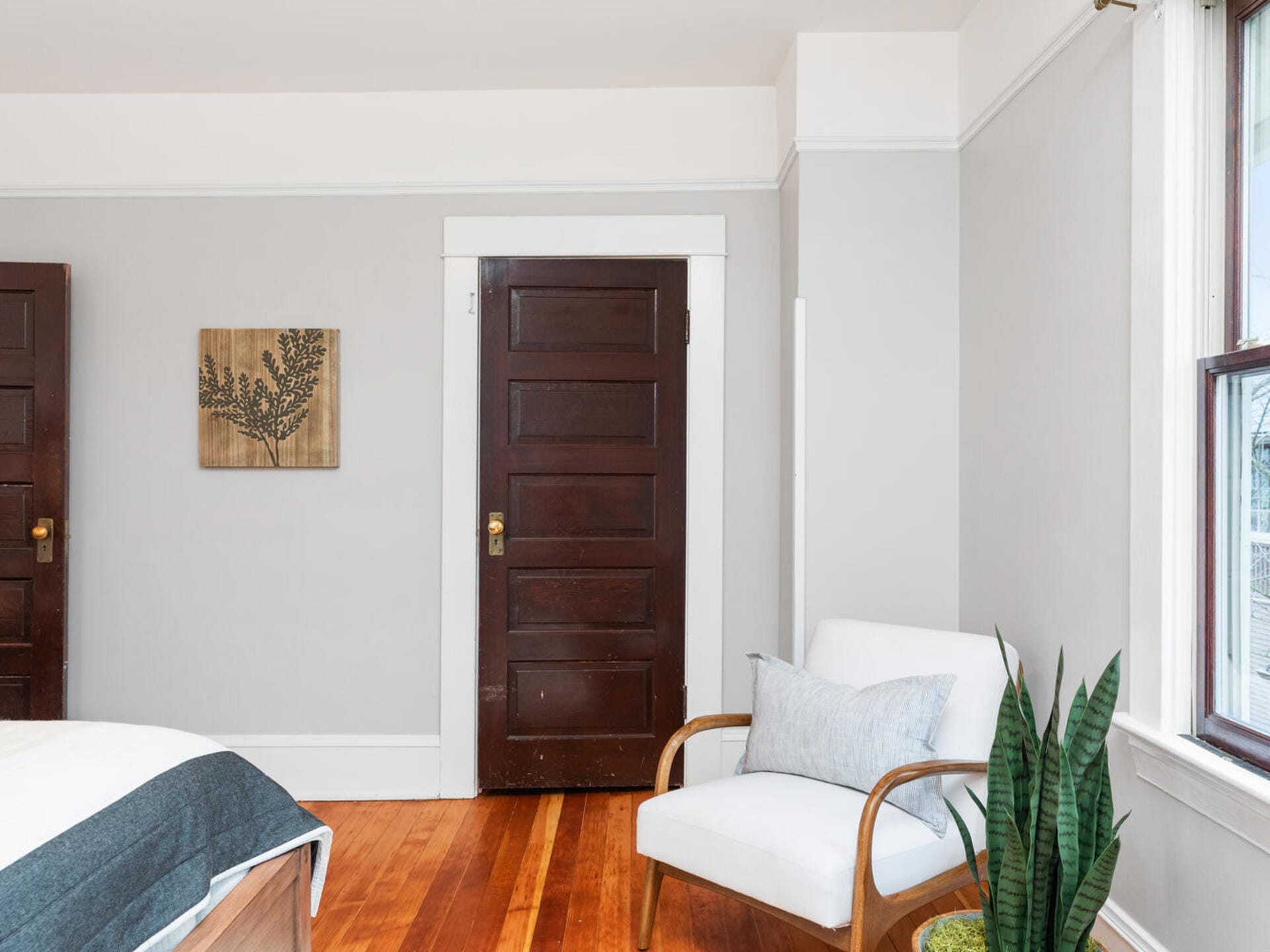 A cozy bedroom corner with a white armchair and a green plant beside it. Dark wooden doors stand against light gray walls. A wooden wall art piece featuring a leaf design is hung on the wall. Sunlight streams through a nearby window.