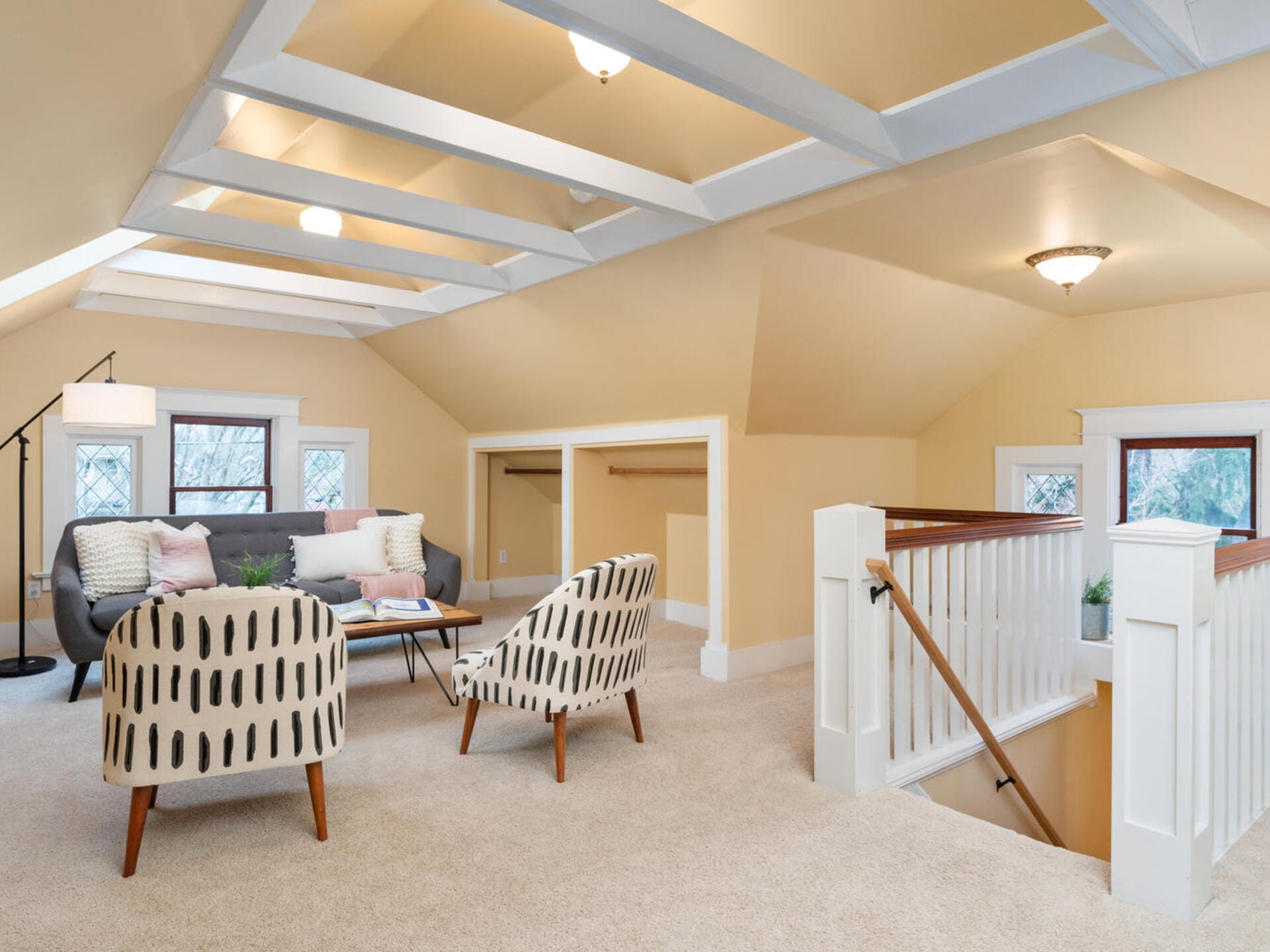 A cozy attic living space with beige walls and carpet. It features a gray sofa, two patterned armchairs, a wooden coffee table, and a floor lamp. Skylights and small windows provide natural light, and a staircase with white railing leads downstairs.