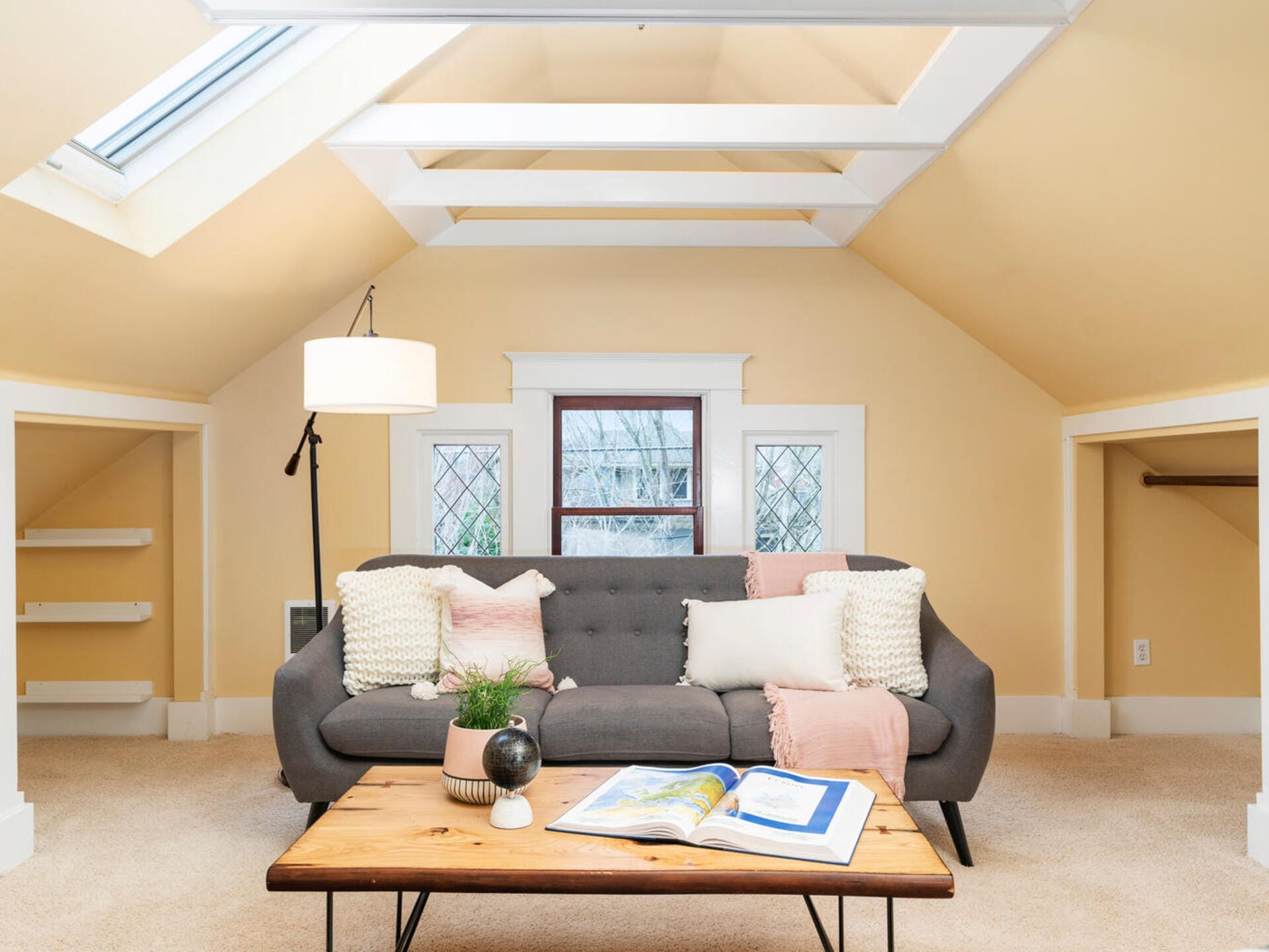 A cozy attic living space with a gray sofa adorned with pink and white cushions. A wooden coffee table holds a plant and an open book. Soft yellow walls, a floor lamp, bookshelves, and a skylight brighten the room.