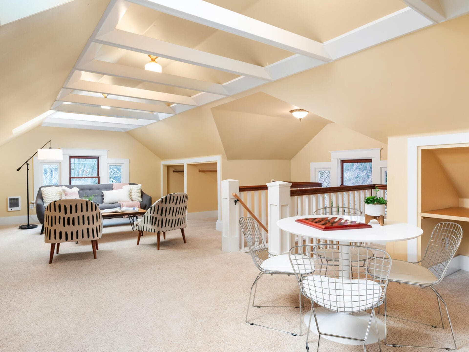 A spacious attic living area with cream walls and carpet, featuring a seating area with a gray sofa and patterned chairs, a white round dining table with wire chairs, and skylights. The space is modern and well-lit with a cozy ambiance.