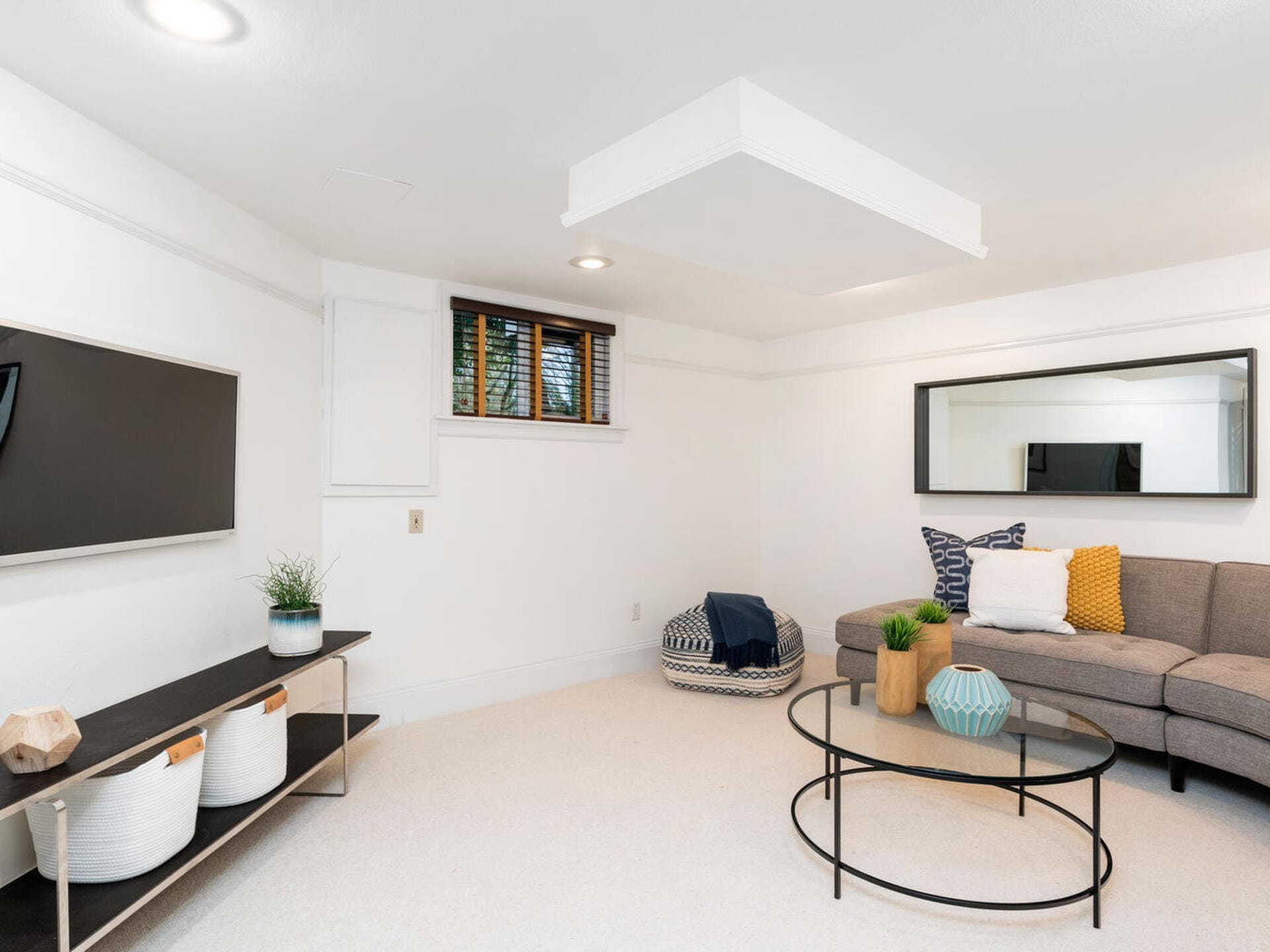 A modern living room with a gray sectional sofa, decorative pillows, and a round glass coffee table. A wall-mounted TV is on the left, below which is a console table with plants and baskets. A large mirror is on the wall above the sofa.