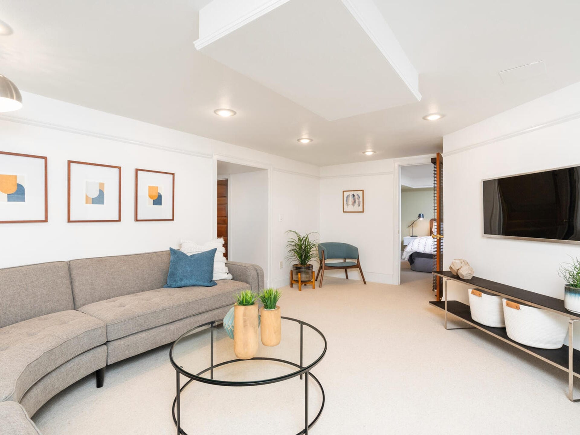A modern living room with a gray sectional sofa, round glass coffee table with decorative vases, wall art, and a flat-screen TV. The room is spacious and bright, featuring a chair and plant in the corner and a view into a bedroom through an open doorway.