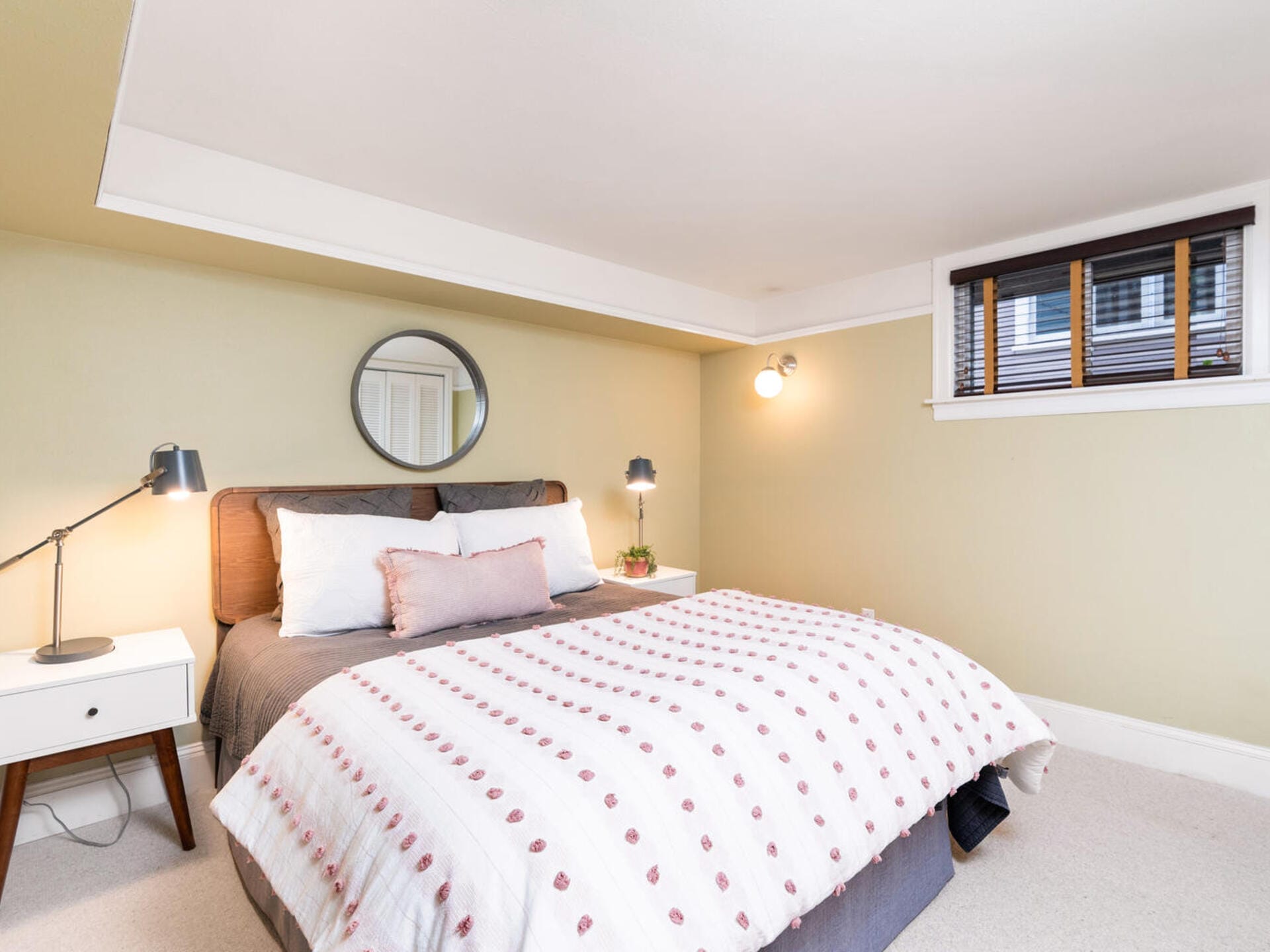 Cozy bedroom with a large bed featuring a polka dot comforter, flanked by two bedside tables with lamps. A round mirror hangs above the bed on a light green wall. A small window with blinds is partially open, letting in soft light.