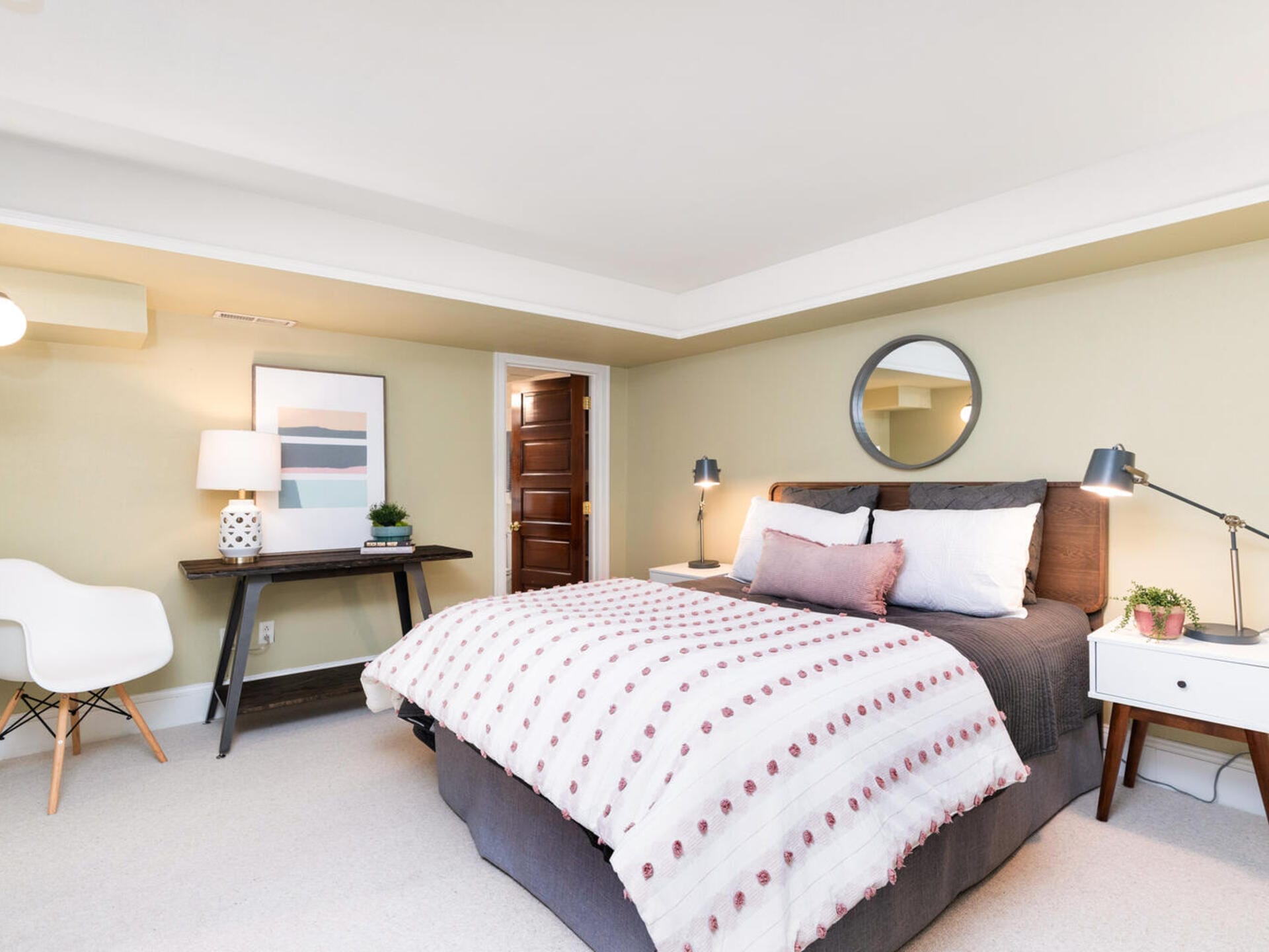 A cozy bedroom with a bed adorned with patterned bedding and pillows. Two side tables with lamps flank the bed. A circular mirror hangs above the bed. Theres a dark wooden desk with a lamp and decor items on the left. Soft lighting brightens the room.