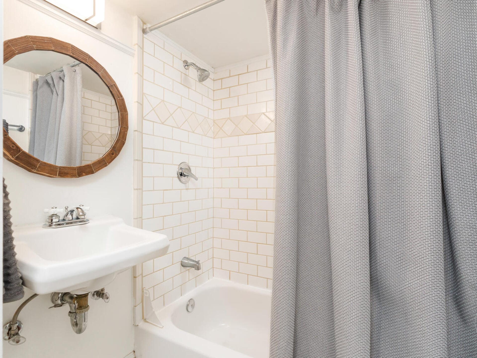 A small bathroom with a white tiled wall and bathtub, grey shower curtain, white sink with a single faucet, and a circular mirror with a wooden frame above the sink. The floor and countertop are not visible.