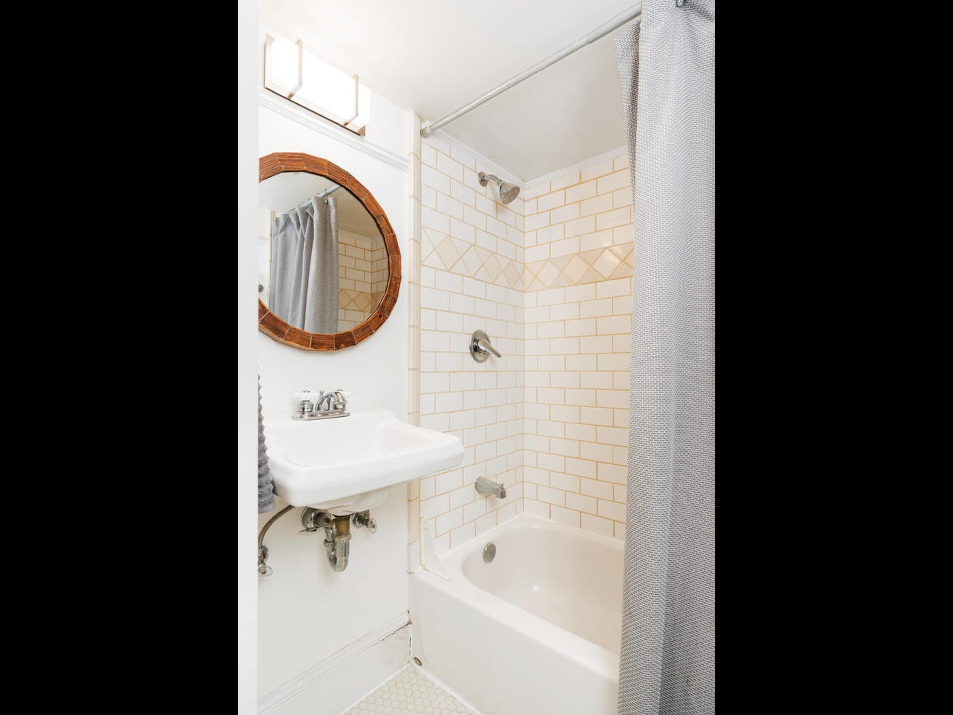 Small bathroom with a white-tiled bathtub and shower, featuring a gray curtain. A round mirror with a wooden frame hangs above a white sink. The floor is tiled in a light pattern, and there is a towel hanging beside the sink.