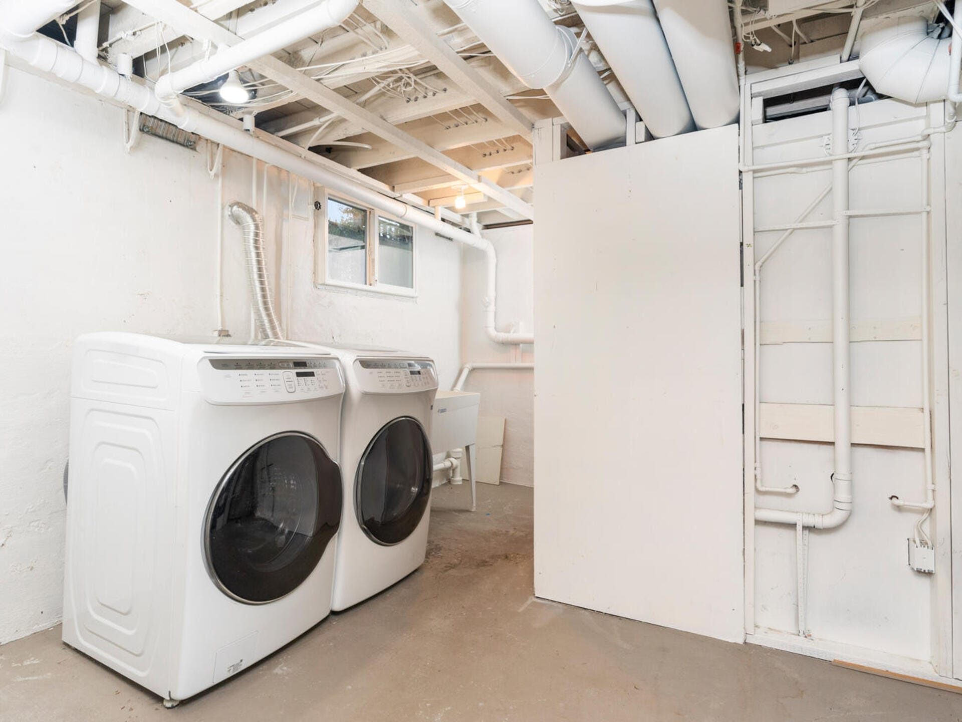 A basement laundry area with a washer and dryer set side by side against a white concrete wall. Exposed pipes run along the ceiling, and a small window provides natural light. A utility sink is also visible.