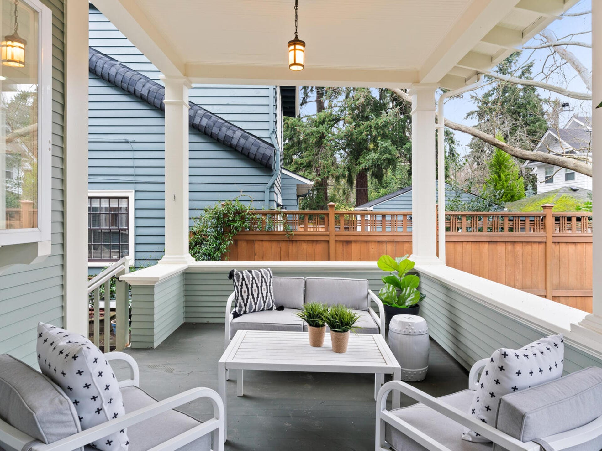 Covered porch with white ceiling and hanging light, featuring gray cushioned seating and a white coffee table with potted plants. Surrounded by a wooden railing and adjacent to a blue house. Trees and a wooden fence are visible in the background.