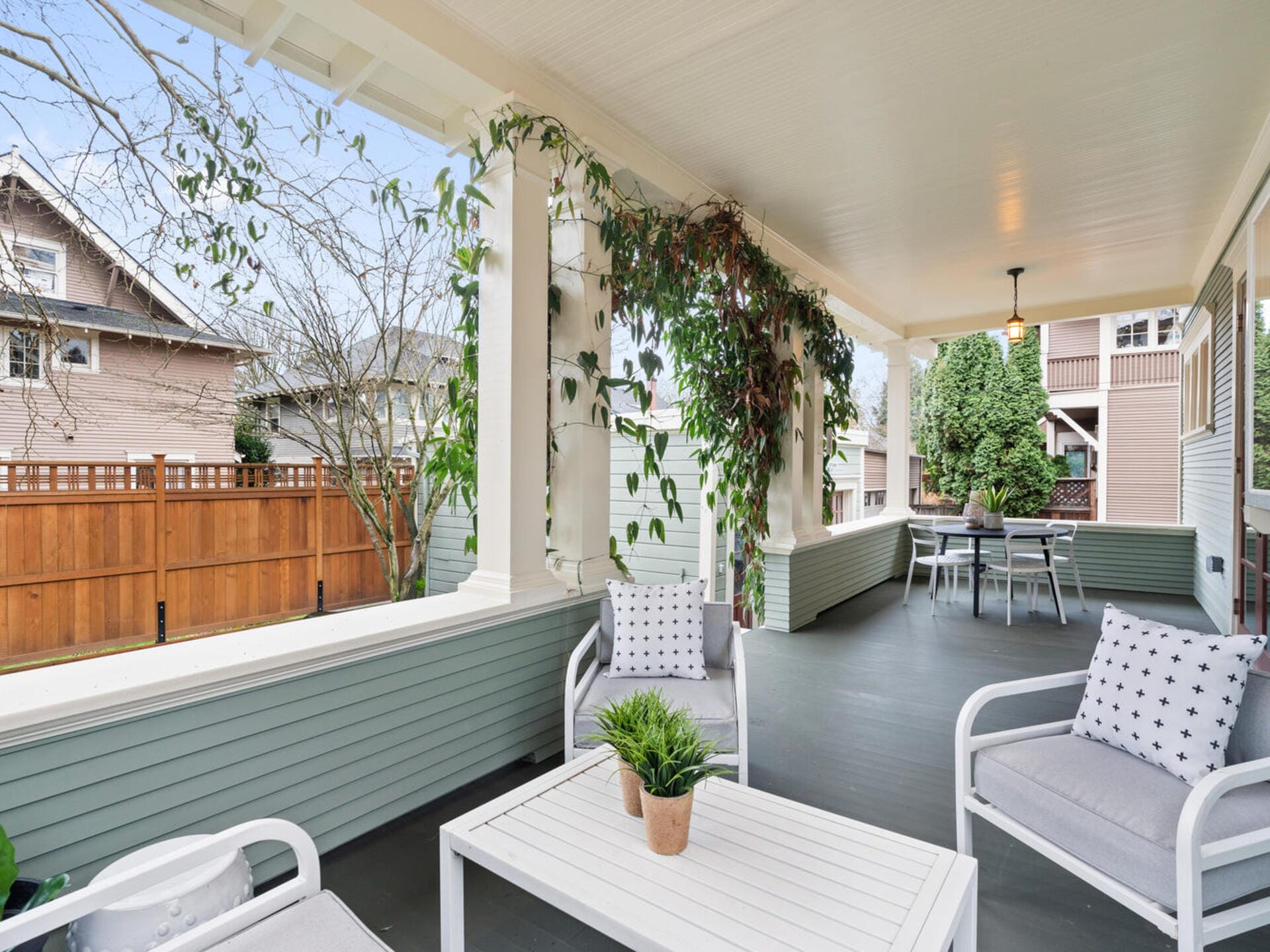 A cozy porch with white furniture and potted plants. The seating area includes cushioned chairs and a coffee table adorned with a small plant. Vines climb the porch columns, and a wooden fence and neighboring houses are visible in the background.