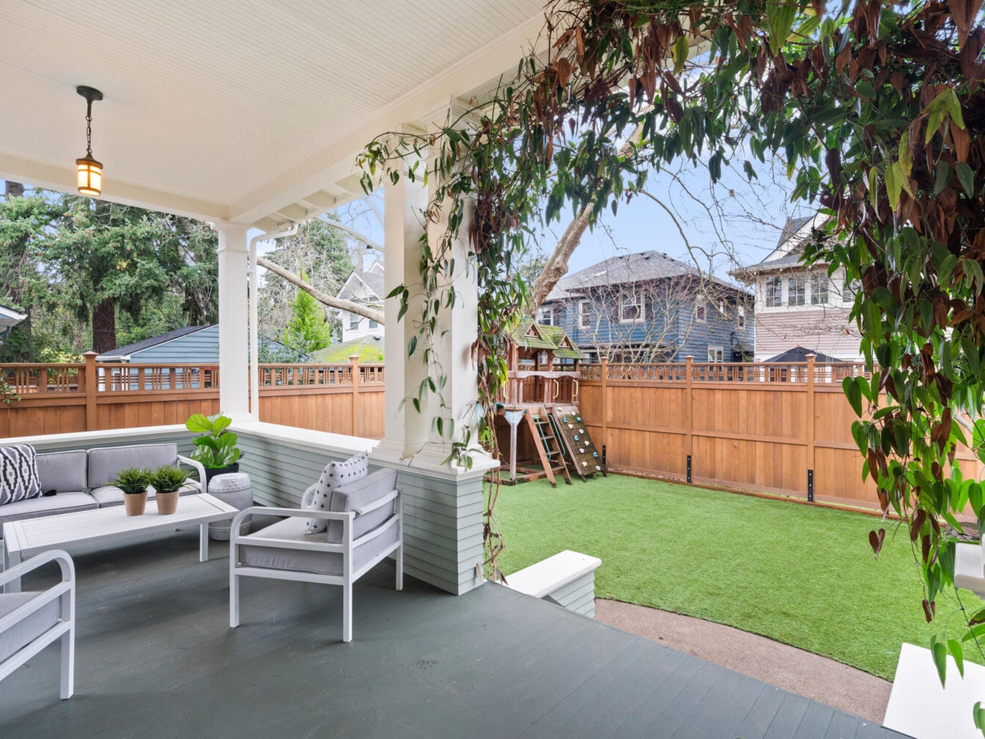 A cozy covered patio features a white outdoor sofa and chairs with cushions, a potted plant, and overhead lighting. Climbing plants adorn the beams. Beyond the wooden fence, a grassy backyard with a wooden playground set is visible.