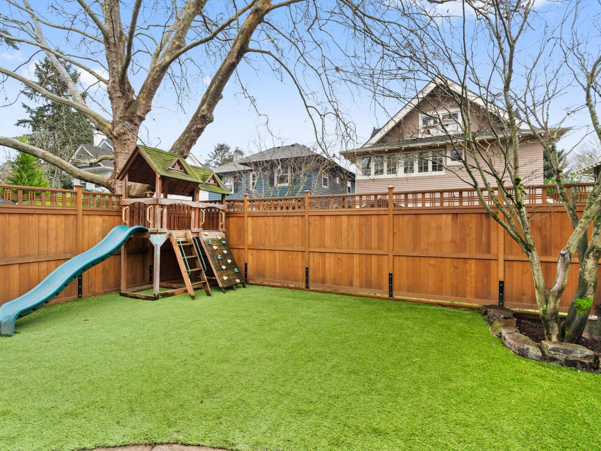 A backyard with artificial grass features a wooden playset, including a slide and climbing wall. A large tree provides partial shade. A tall wooden fence surrounds the yard, and two houses are visible in the background.