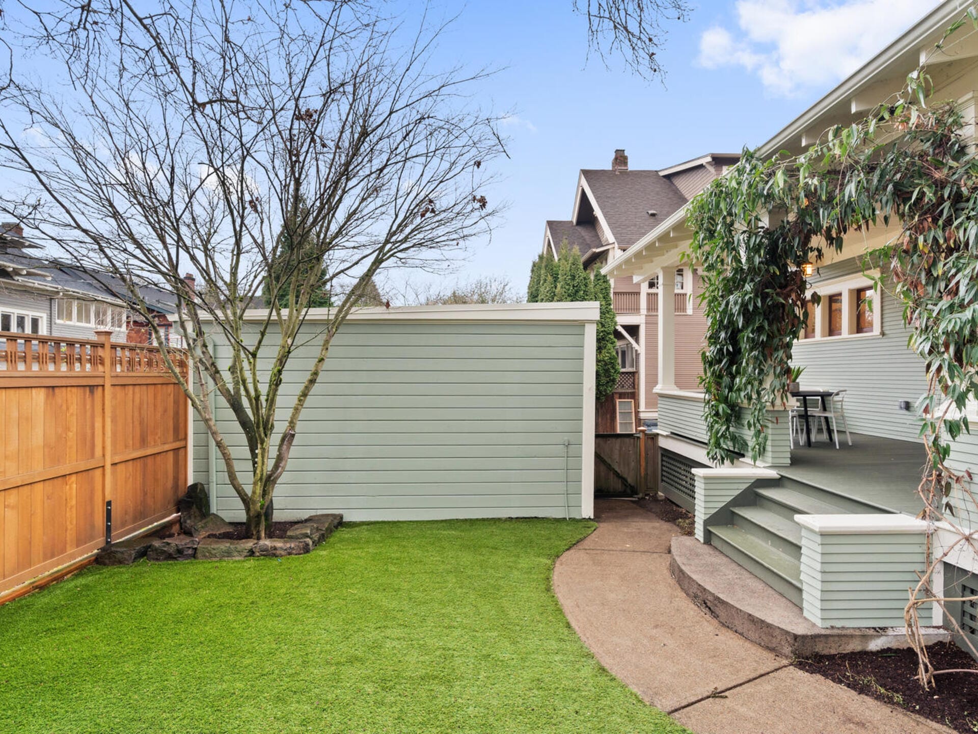 A backyard with a small green lawn, a leafless tree, and a wooden privacy fence. The yard features a green shed and a covered patio with steps and a hanging plant, beside a house with beige siding and a gabled roof.