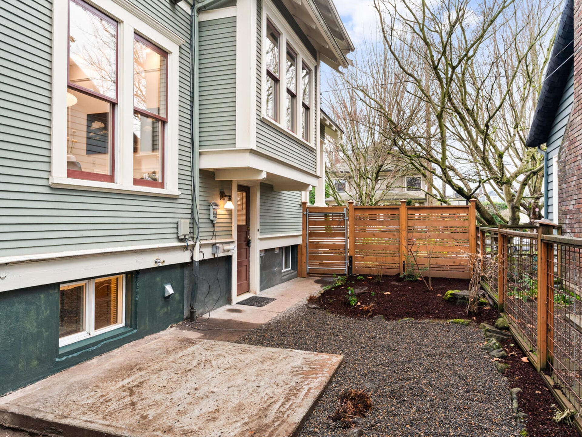 Small backyard with a gravel area and concrete path next to a green, two-story house. There’s a wooden privacy fence on the right, bare trees, and large windows on the house. The sky is partly cloudy.
