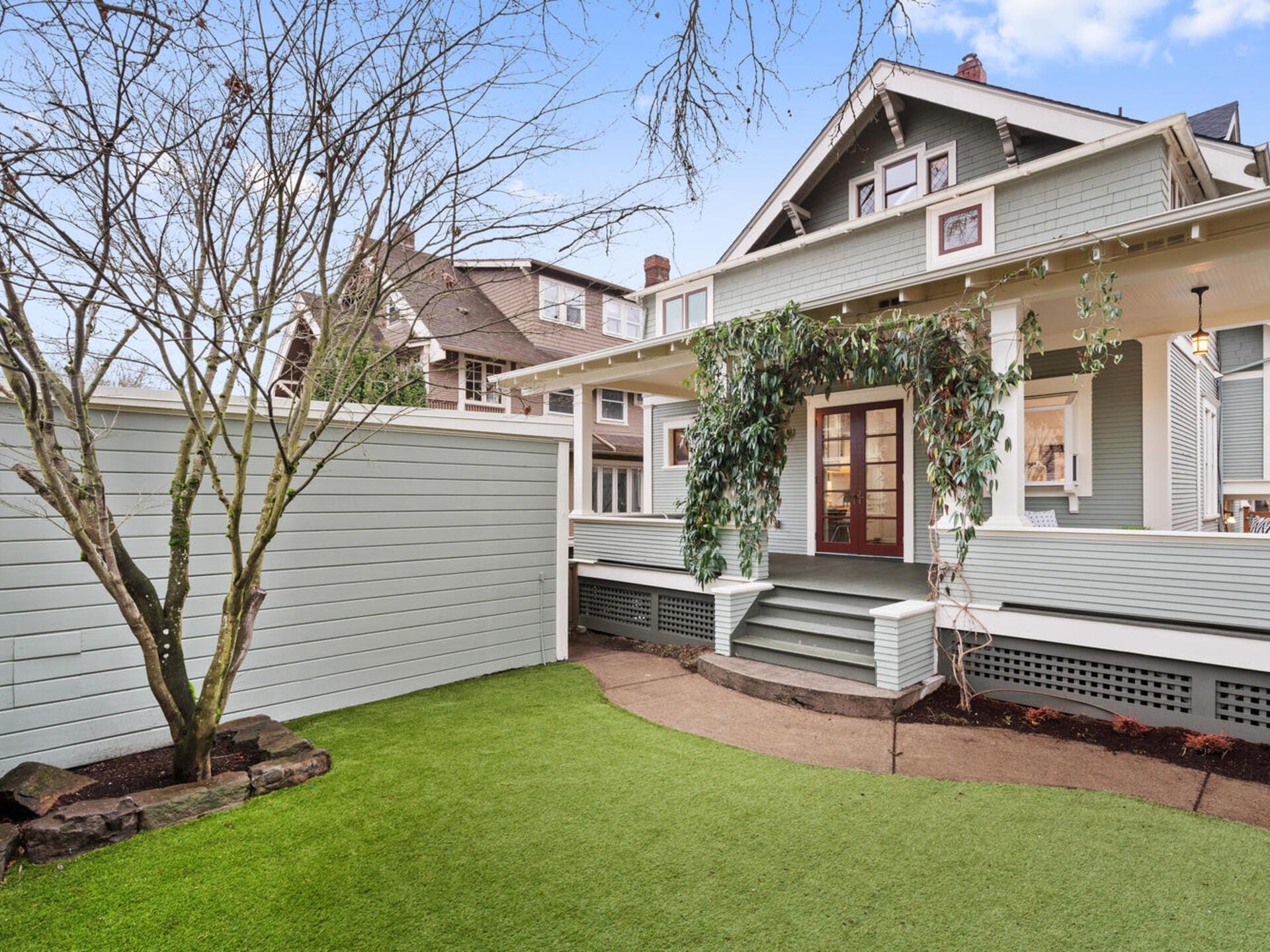 A charming, light green house with a large porch and overhanging plants. It features a manicured artificial lawn and a surrounding white fence. The sky is clear and blue, adding to the serene atmosphere.
