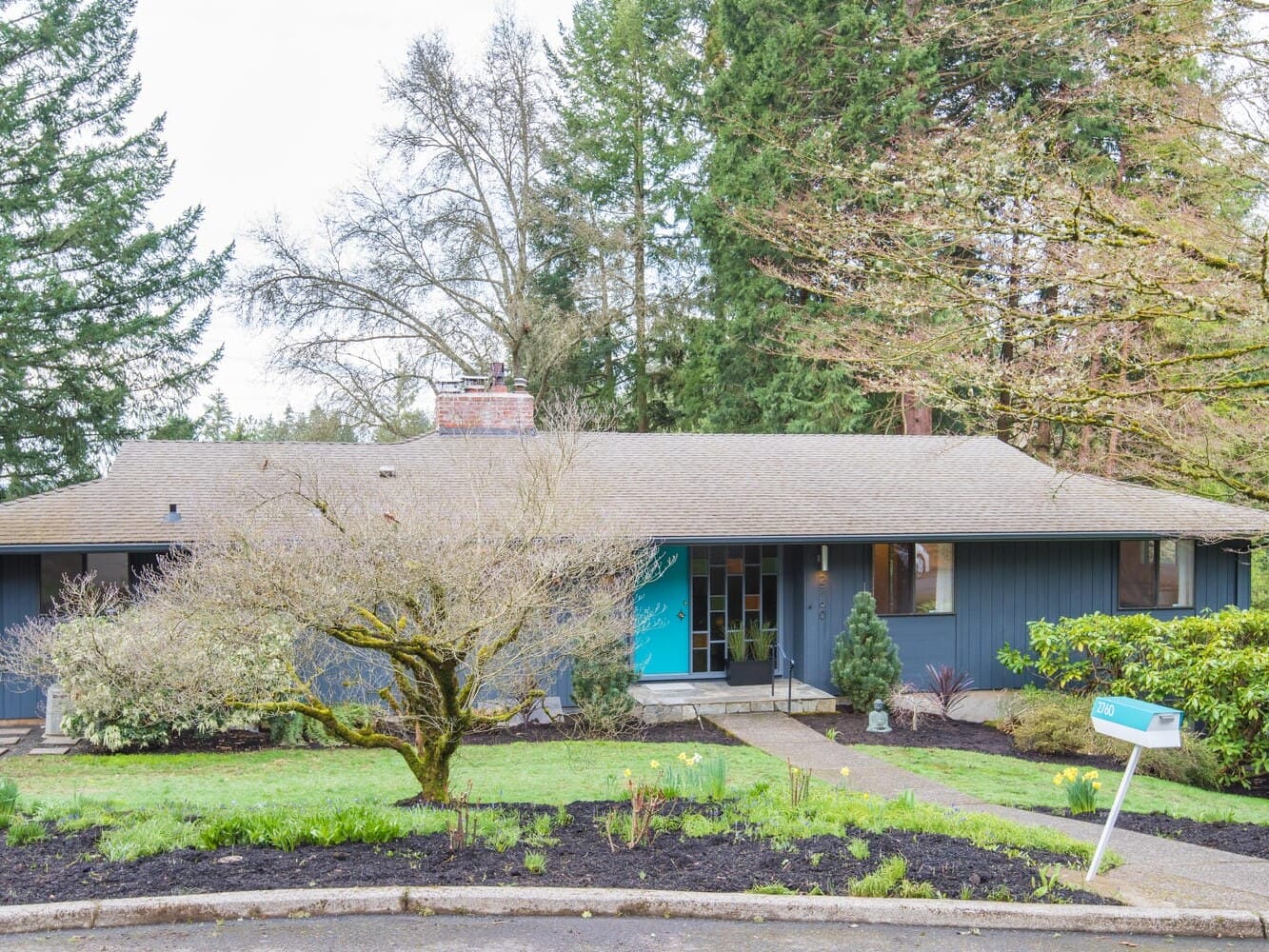 A mid-century modern house in Portland, Oregon, features a blue facade and brick chimney. It boasts a turquoise door, large windows, and a well-maintained front yard with trees and shrubs. A mailbox stands proudly by the street as tall trees cocoon the property in nature's embrace.