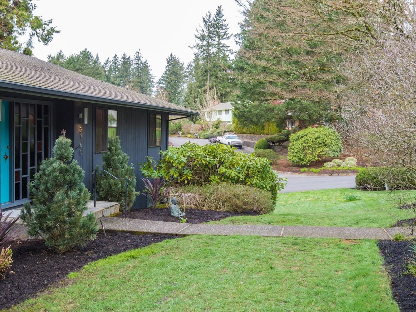 A suburban street in Portland, Oregon, showcases a blue house on the left with a glass front door and trimmed shrubs. The sidewalk leads to a quiet road lined with trees and houses. It's a cloudy day with lush greenery enveloping the area.