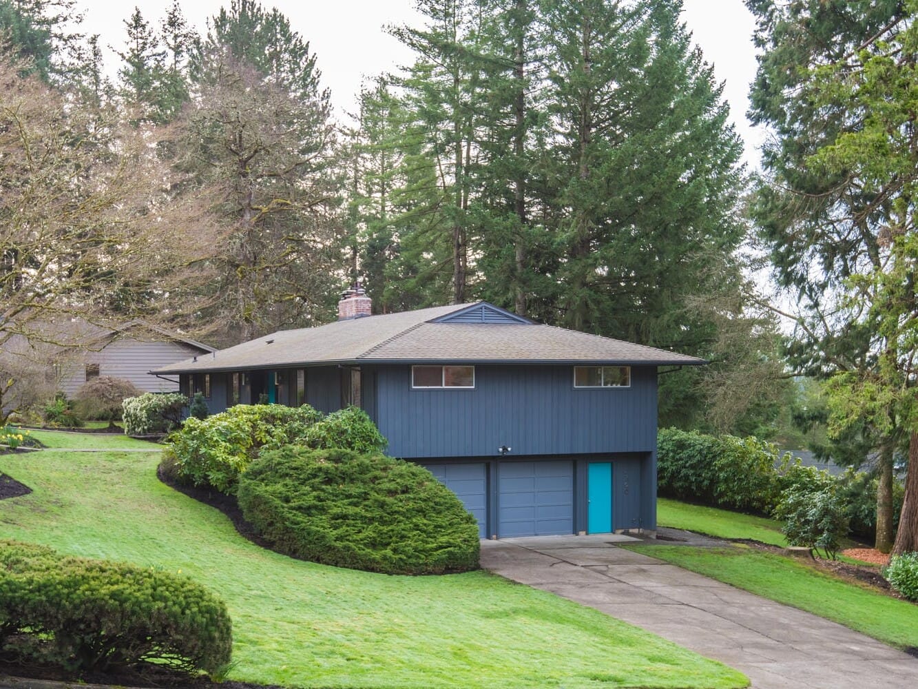 A modern blue-gray house in Portland, Oregon features a double garage and is surrounded by a lush green lawn and tall trees. The driveway gracefully curves up to the house, while the yard is beautifully landscaped with neatly trimmed bushes.