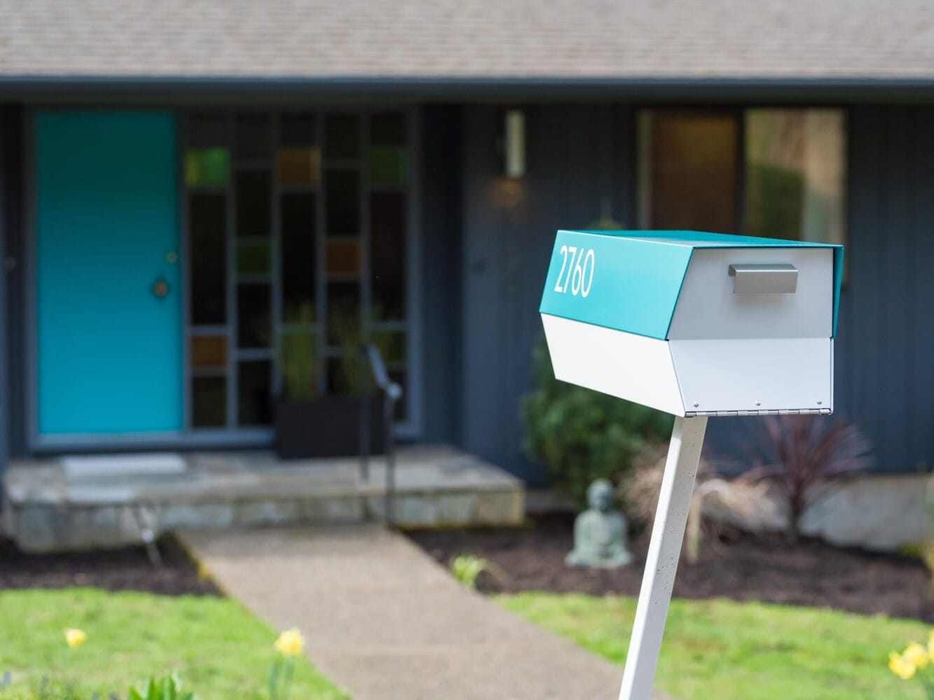 A modern house in Portland, Oregon, features a blue door and an abstract window design. In the foreground, a teal and white mailbox bears the number 2160, beside a stone path lined with vibrant yellow flowers.