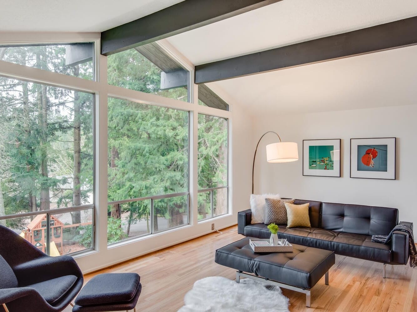 Bright living room with large windows overlooking Portland, Oregon's lush trees. Features a black sofa, modern chair, floor lamp, and coffee table on hardwood flooring. Two colorful art pieces hang on the wall. A fluffy white rug adds texture.