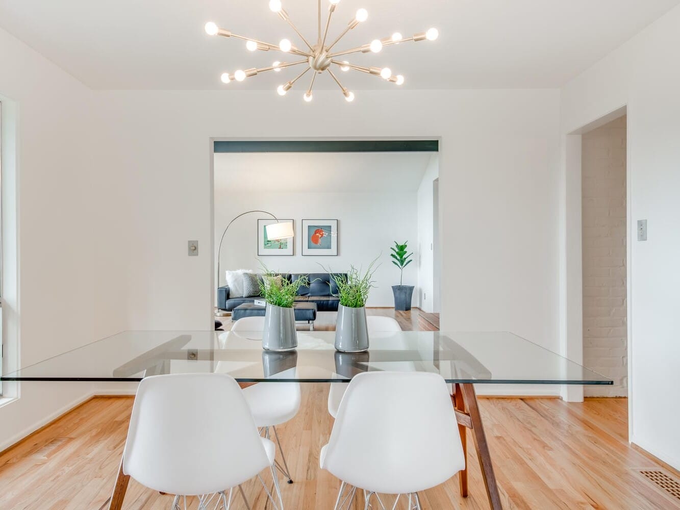 A modern dining room in Portland, Oregon, features a glass table, four white chairs, and two potted plants. A contemporary chandelier hangs above. The room boasts wooden floors, while in the background, a living area adorned with wall art and an additional potted plant is visible.