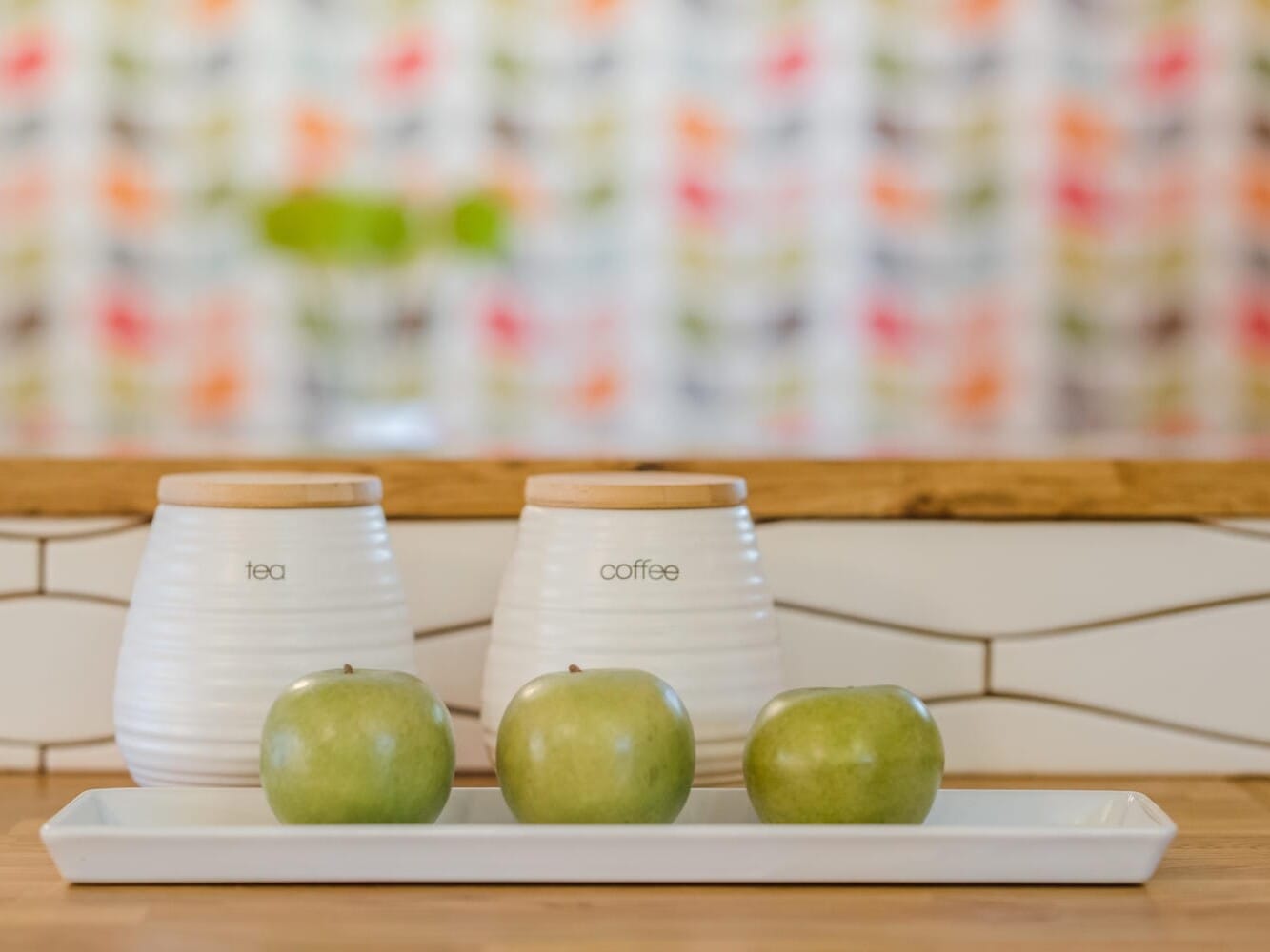 A kitchen counter in Portland, Oregon displays two white containers labeled tea and coffee, topped with wooden lids. In front, a rectangular white tray holds three green apples. The background showcases a colorful, blurred pattern.