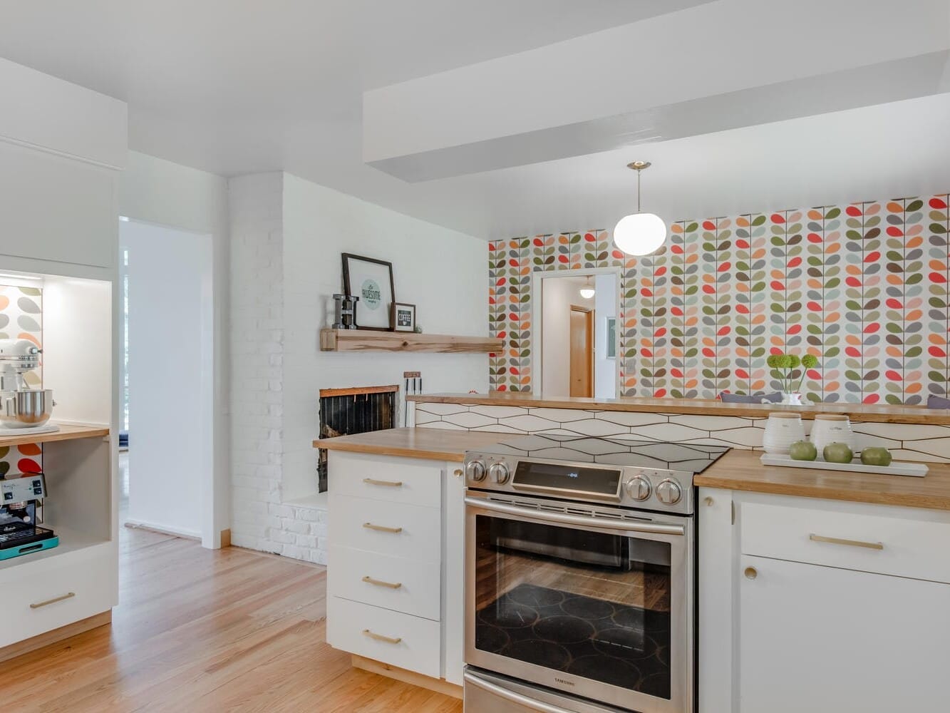 A modern kitchen in Portland, Oregon, with a cozy, colorful design. It features a sleek stainless steel stove, light wood countertops, and white cabinetry. The vibrant patterned wallpaper on the walls is complemented by wooden flooring and decorative elements on a shelf.