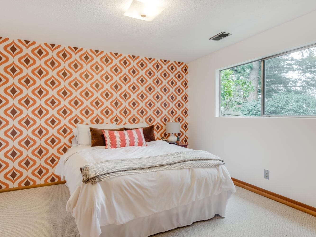 A bedroom with a patterned orange and white accent wall behind a neatly made bed adorned with pillows and a throw blanket. A large window on the right allows natural light, revealing greenery outside typical of Portland, Oregon. The room features beige carpet and white walls.