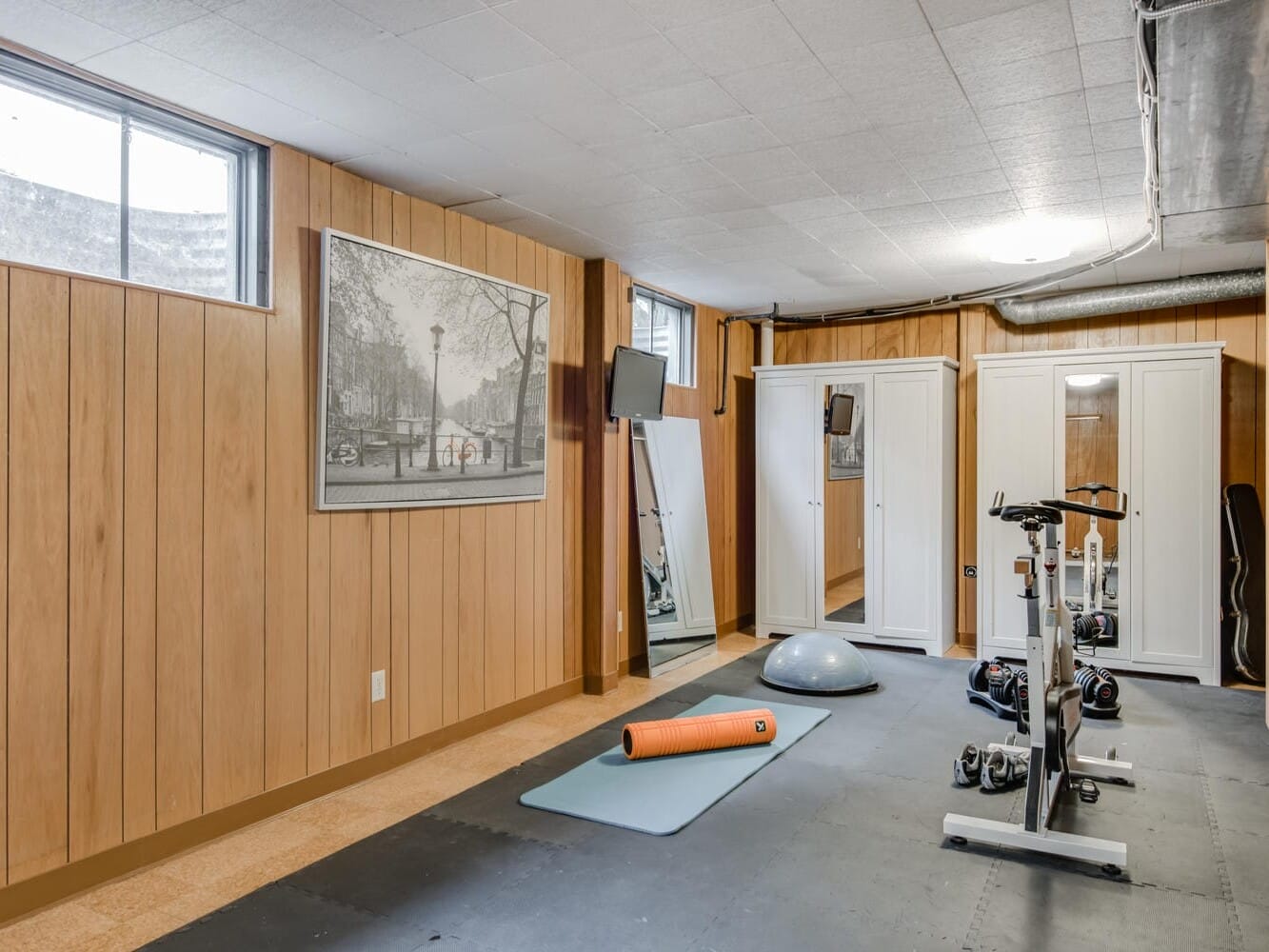 A home gym setup in a Portland, Oregon basement features an exercise bike, yoga mat, balance trainer, and foam roller on rubber flooring. Light wood-paneled walls display a large cityscape photo, and mirrors are seen on the wall. A small window lets in natural light.