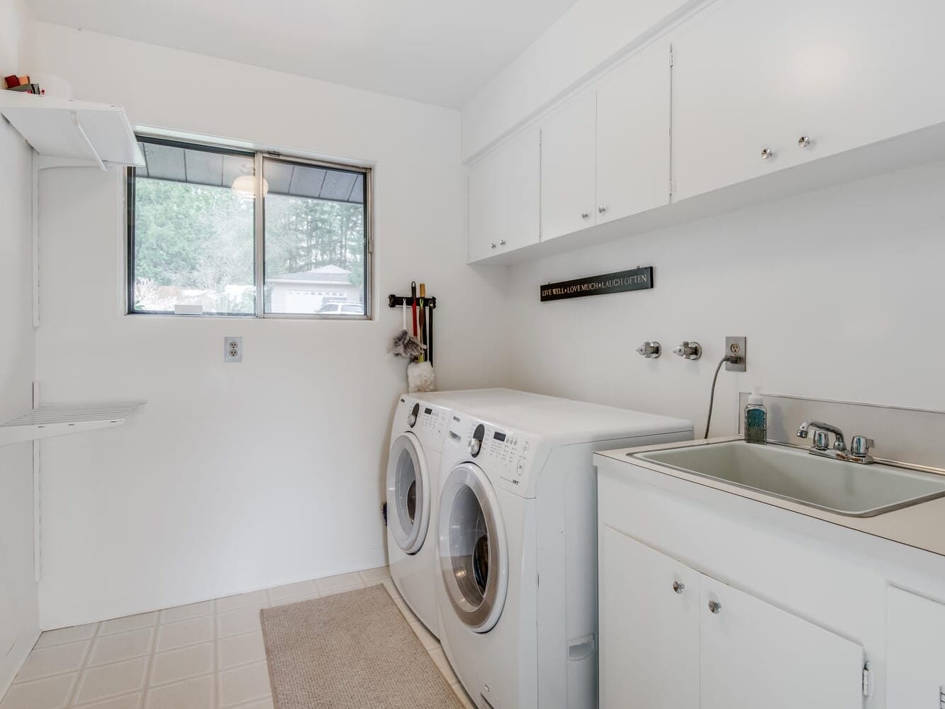 A tidy Portland, Oregon laundry room features a washer and dryer set, a utility sink, and white cabinets above. A window allows natural light in, complemented by a small shelf mounted on the left wall.