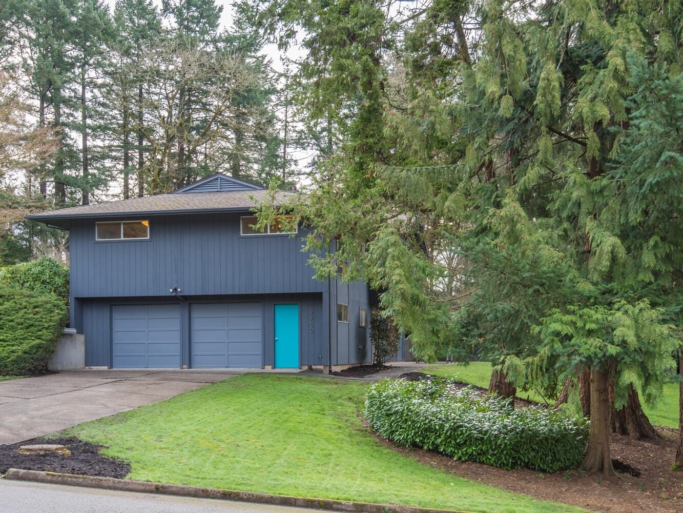 A two-story blue house in Portland, Oregon, with a teal front door is partially obscured by trees. The house features a double garage and is surrounded by green lawns and shrubbery. The scene is peaceful, with tall evergreen trees in the background.