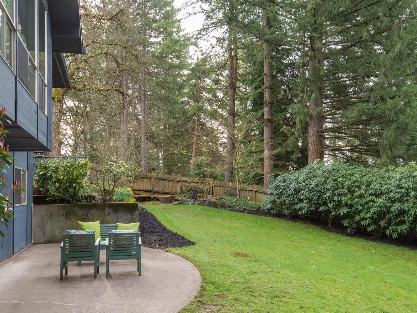 A backyard featuring two green chairs on a concrete patio beside a blue house. The area is surrounded by lush green grass, tall trees, and dense shrubs, creating a serene forest-like setting.