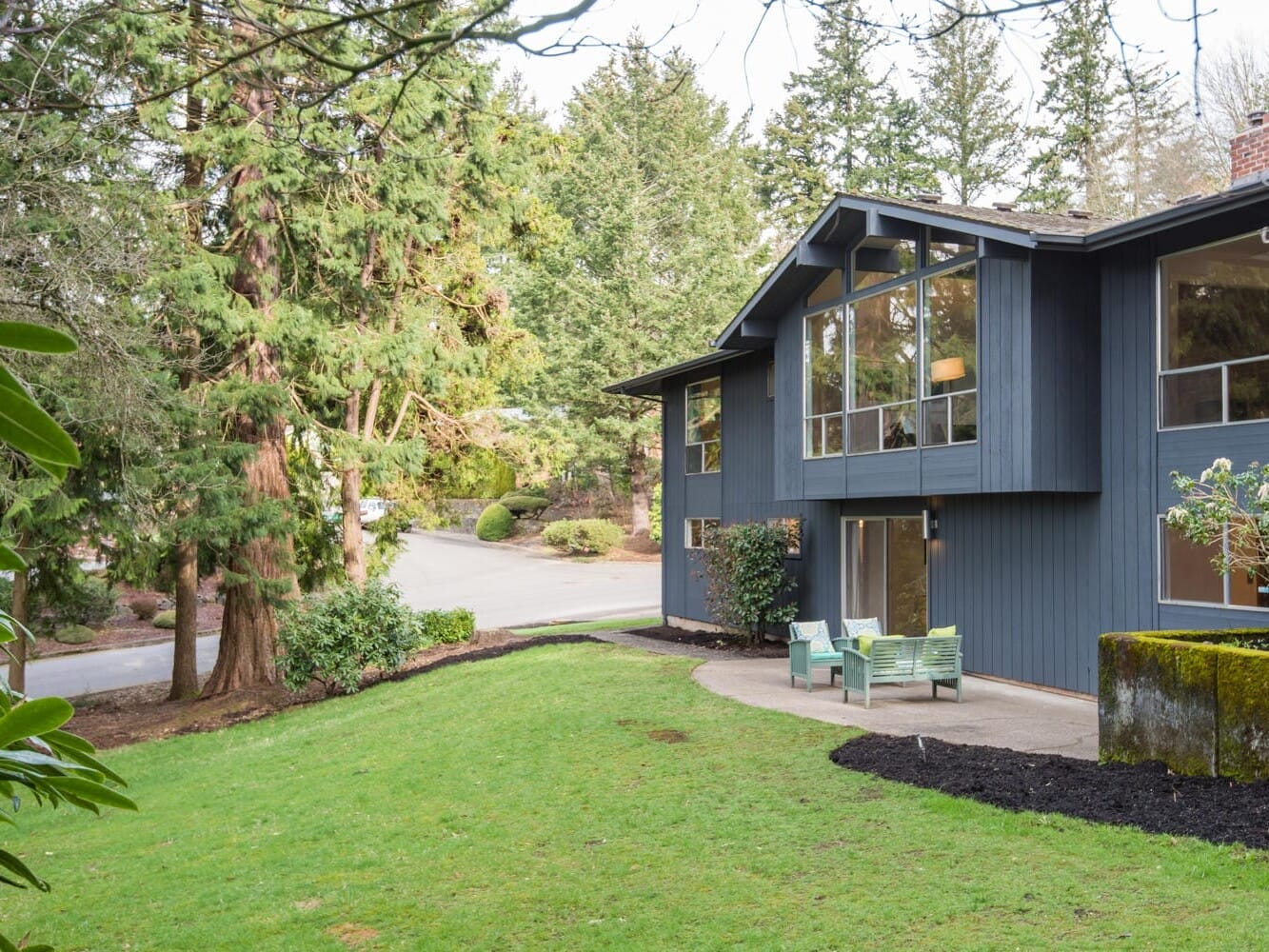 A modern blue house with large windows is surrounded by lush green grass and tall trees. A paved walkway leads to the entrance, while a small patio with a bench is visible in the foreground. The background features a quiet suburban street.