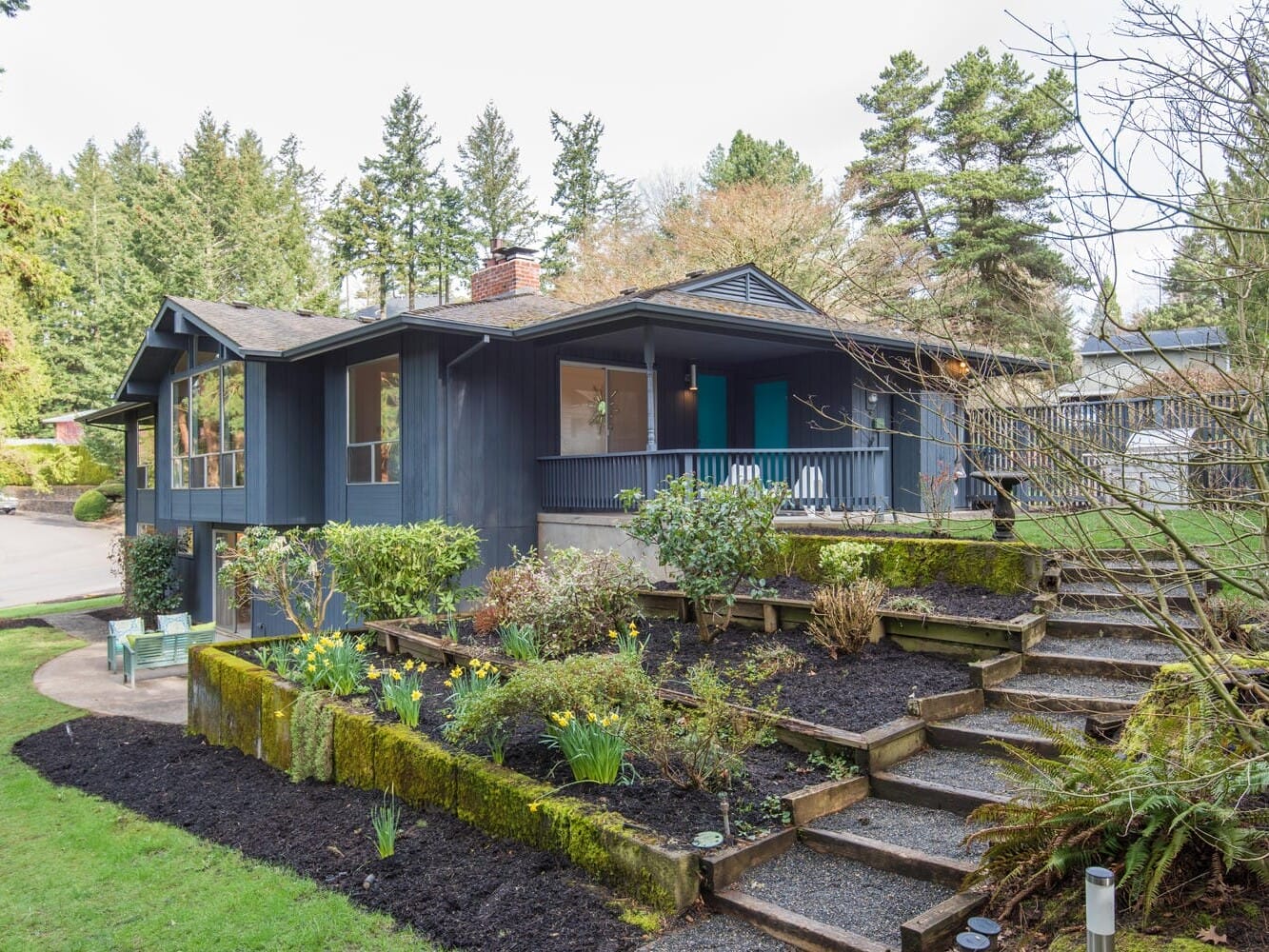 A modern mid-century house with a black exterior surrounded by lush greenery and trees. The front yard features a well-maintained garden with daffodils and a curved stone staircase leading to the porch.