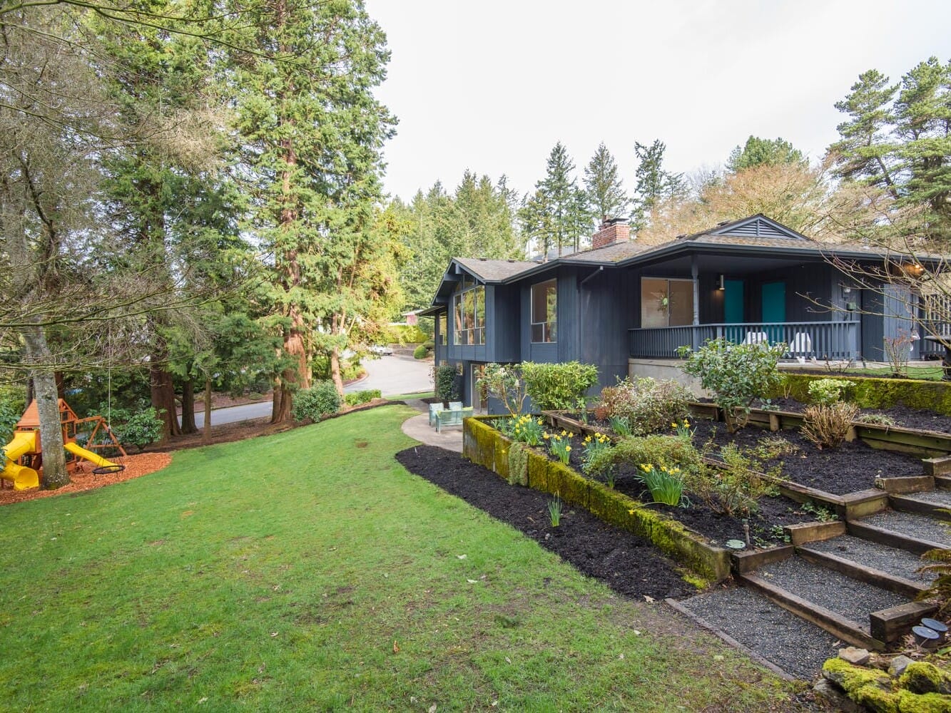 A modern house with a dark exterior is surrounded by lush greenery and tall trees. The sloped yard features well-maintained gardens and a set of stairs leading to the entrance. A play area with a slide is visible among the trees.