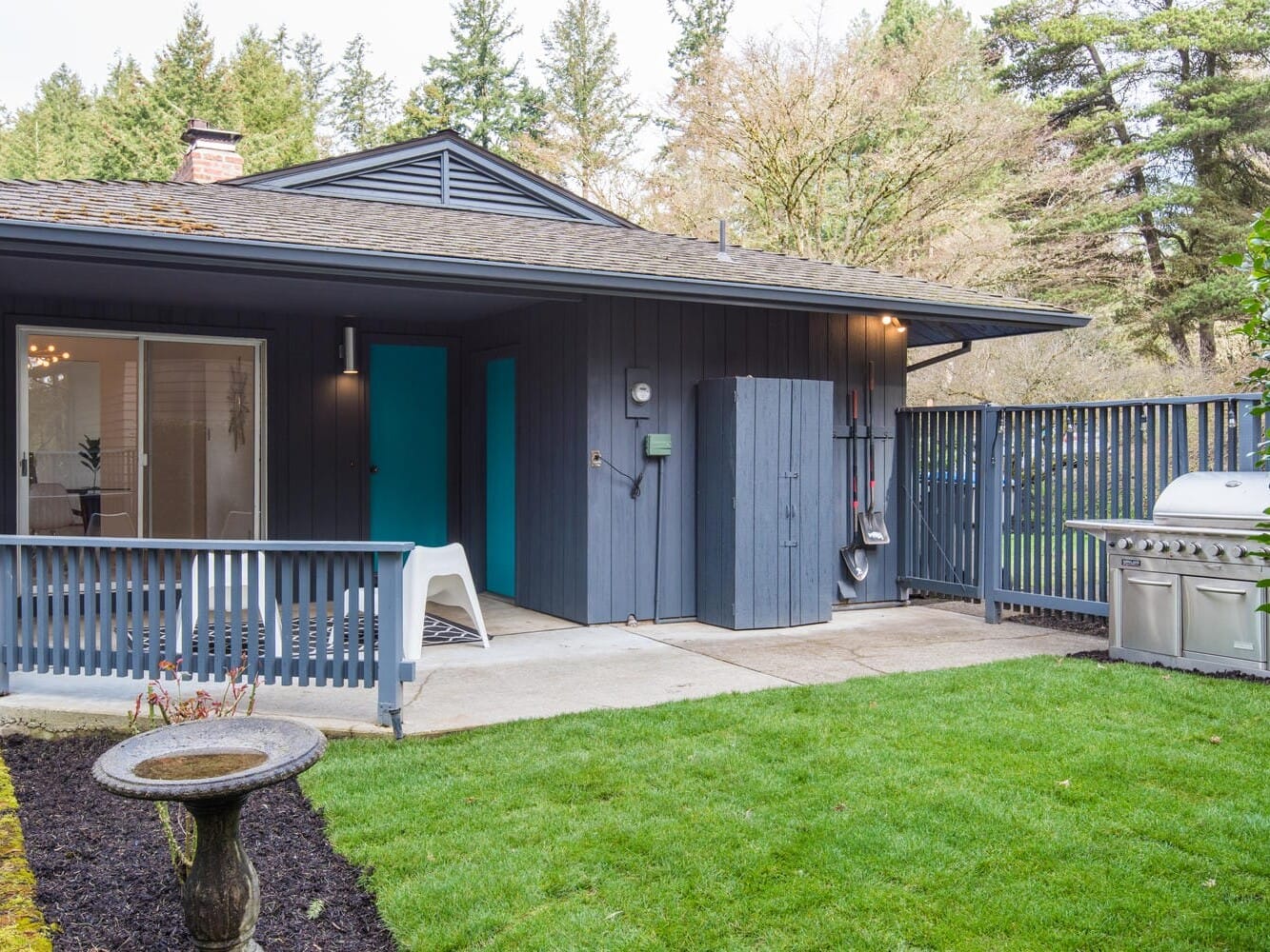 A modern house exterior with gray siding and a turquoise door. Theres a small porch, a birdbath, and a well-kept lawn. A built-in barbecue grill is on a concrete patio, surrounded by a wooden fence, with trees in the background.