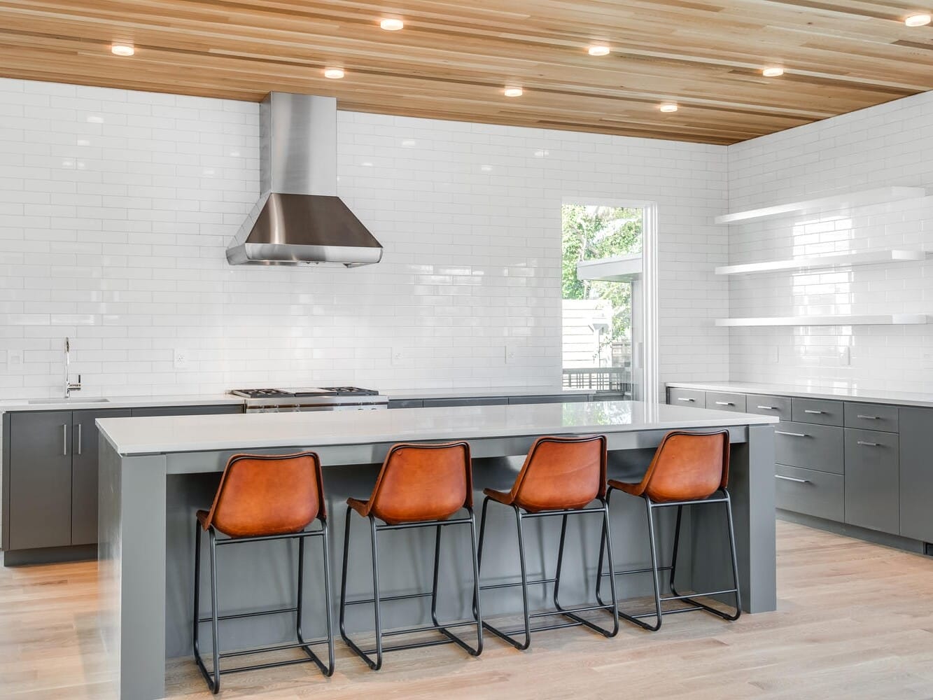 Modern kitchen in Portland, Oregon, showcasing a large island with gray cabinetry and four brown leather bar stools. White subway tile backsplash, stainless steel range hood, and wooden ceiling panels enhance the contemporary design. Natural light fills the space beautifully.