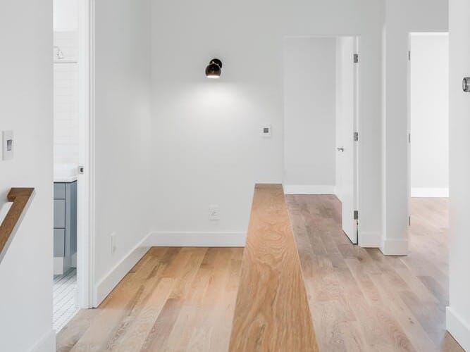 A modern interior hallway in Portland, Oregon, showcases light wood flooring and white walls. The space features a wooden railing and doors leading to adjoining rooms. A single light fixture is mounted on the wall, while natural light floods the area, creating a bright and inviting atmosphere.