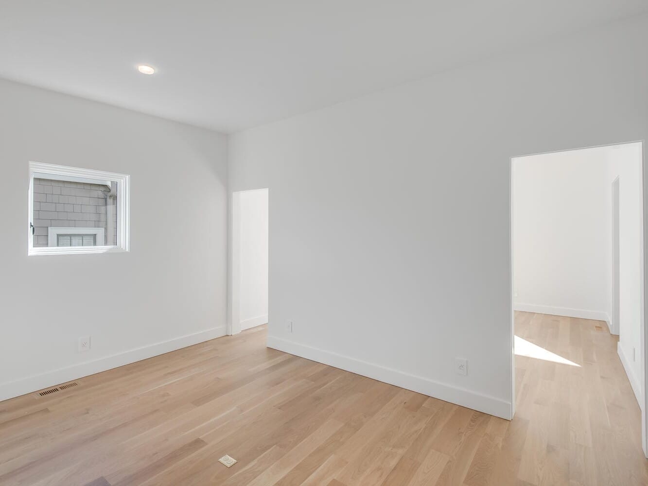 An unfurnished, bright room in Portland, Oregon features light wood flooring, white walls, and a small window. The space leads to another area on the right, also illuminated by natural light.