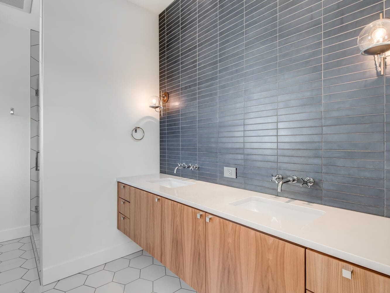 A modern bathroom in Portland, Oregon, showcases a double sink vanity with wooden cabinets, a gray tiled wall, and sleek wall-mounted lights. The white countertop enhances the clean design, while large hexagonal tiles adorn the floor.