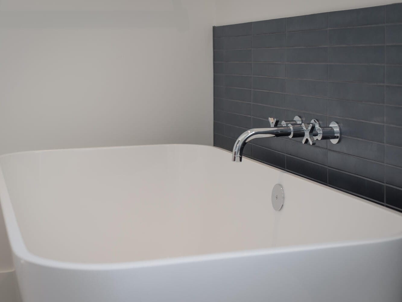 A modern white rectangular bathtub with a shiny silver faucet is set against a wall tiled with dark gray rectangular tiles, creating a minimalist bathroom aesthetic reminiscent of Portland, Oregon's sleek design trends.