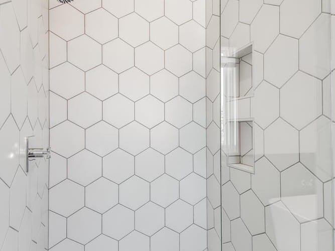 A modern shower in Portland, Oregon, boasts large hexagonal white tiles on the walls and small black hexagonal tiles on the floor. It features a sleek silver showerhead and a recessed wall shelf, all enclosed by elegant glass doors.