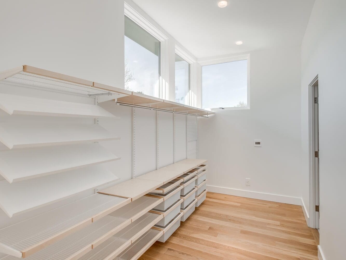 A minimalist room in Portland, Oregon, with a wooden floor, features empty white shelves, drawers, and a pegboard on the left wall. Bright sunlight enters through two large windows above, creating a clean and airy atmosphere.