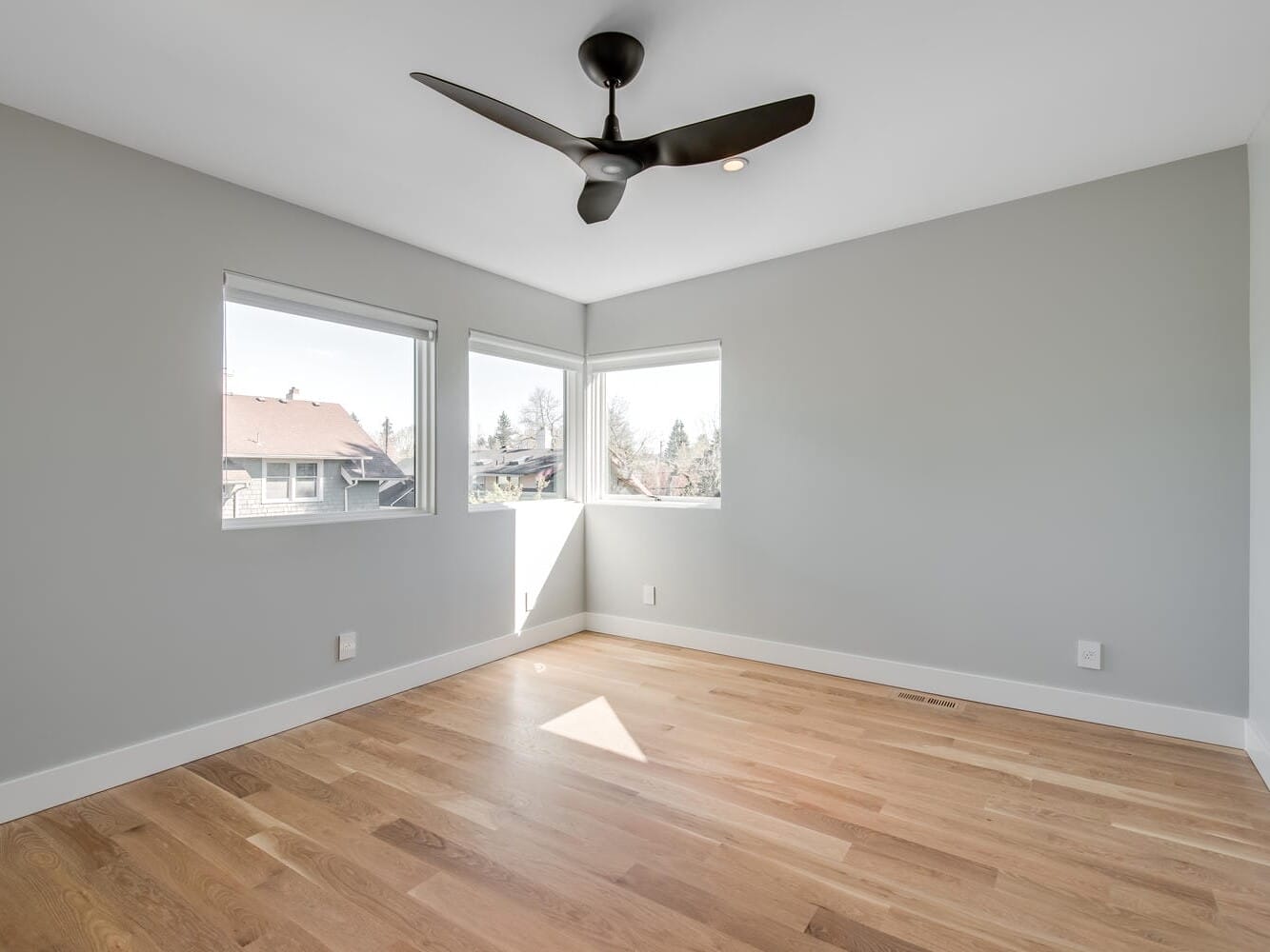 A bright, empty room in Portland, Oregon, features light gray walls and wooden floors. Three windows flood the space with natural light, while a black ceiling fan is elegantly installed above. A closed door is visible on the right side.