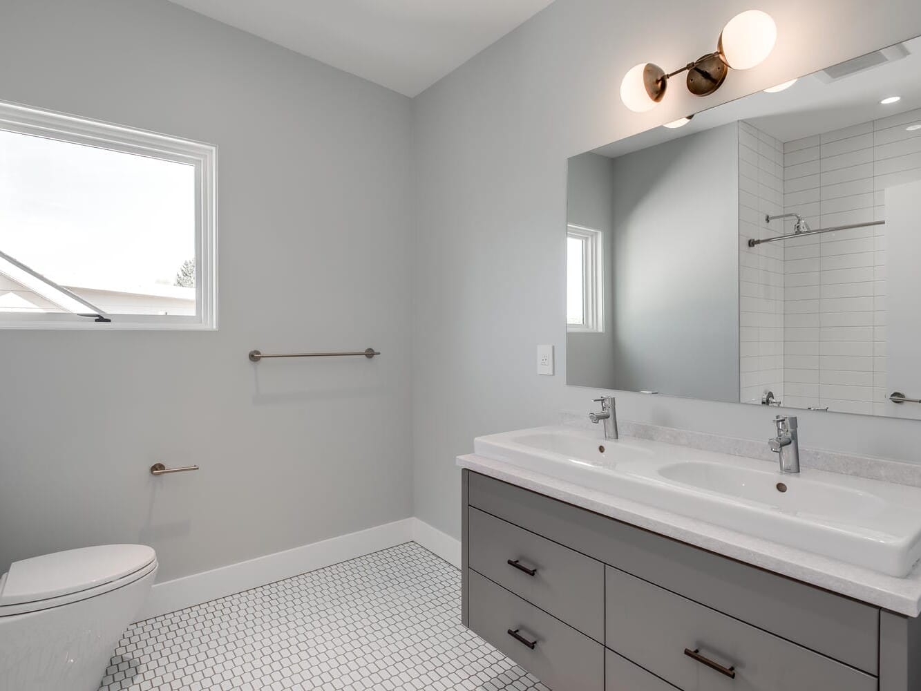 A modern bathroom in Portland, Oregon, features a double sink vanity, a large mirror with wall-mounted lights, and a white toilet. A towel rack sits by a small window. The floor showcases hexagonal tile, while the glass shower enclosure is perfectly reflected in the mirror.