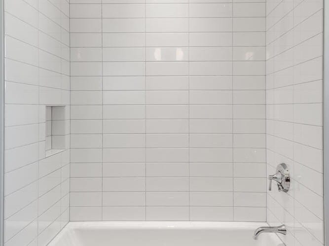 A white tiled bathroom in a Portland, Oregon home features a bathtub with a curved shower rod above. A small recessed shelf adorns the left wall, while a silver showerhead and faucet gleam in the light. The floor showcases a classic pattern of square tiles, adding charm to this cozy space.