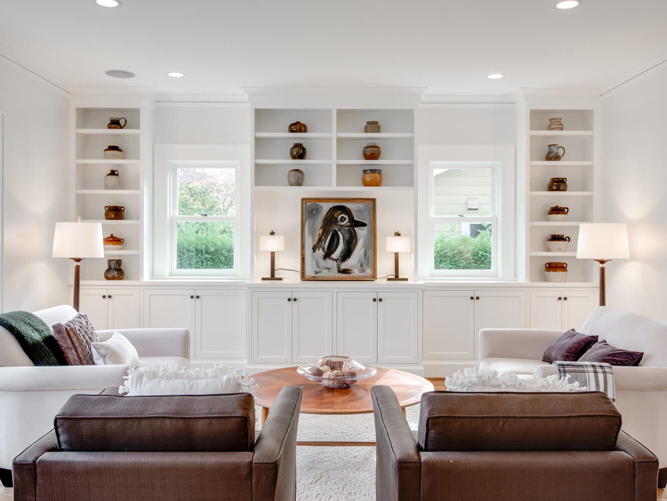 A cozy living room with two white sofas and two brown armchairs around a round wooden coffee table. Built-in shelves with decorative items and a painting are on the wall, flanked by two windows. Lamps provide soft lighting.